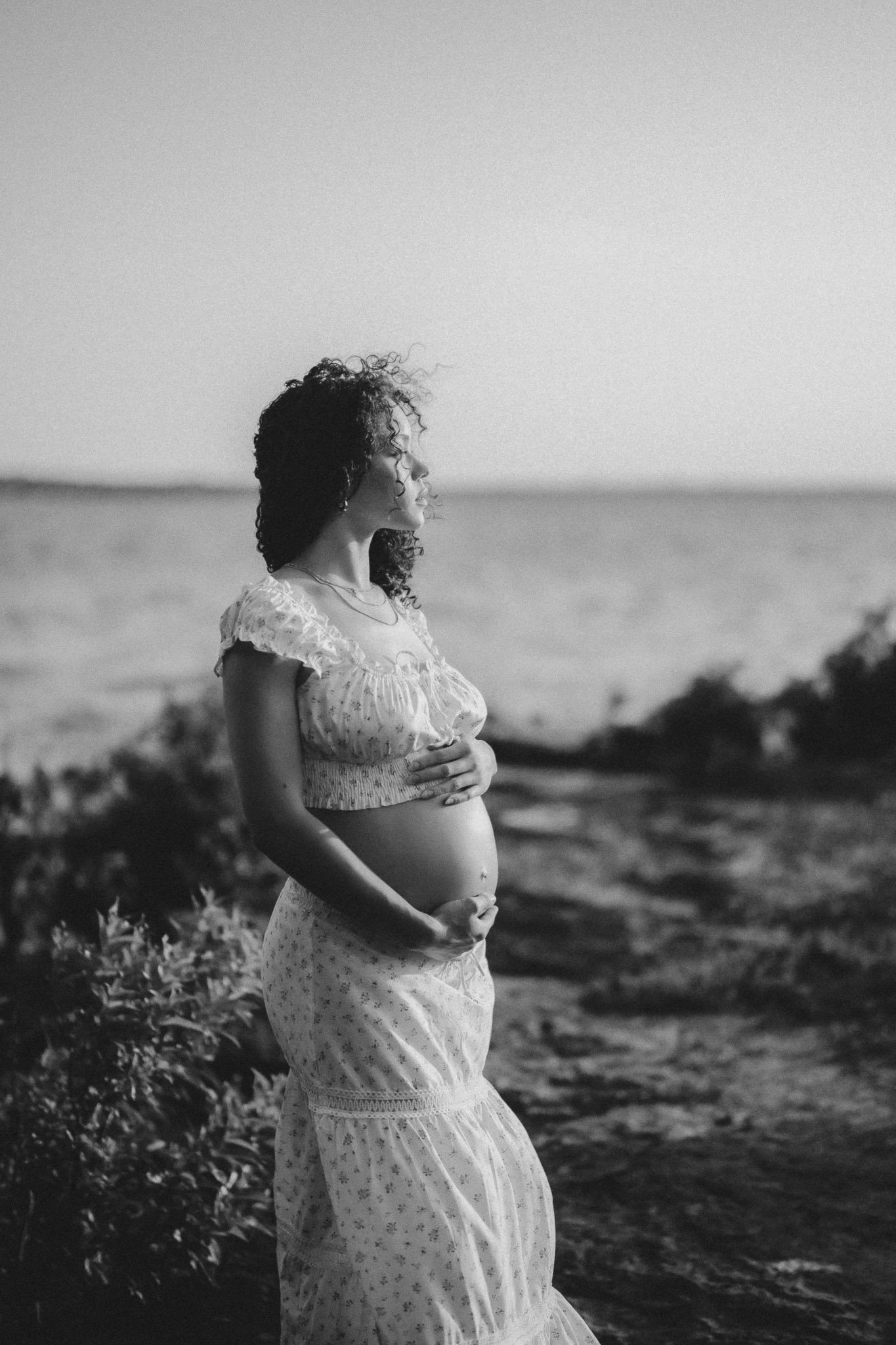 A pregnant woman with curly hair standing on a beach, holding her belly, in black and white.
