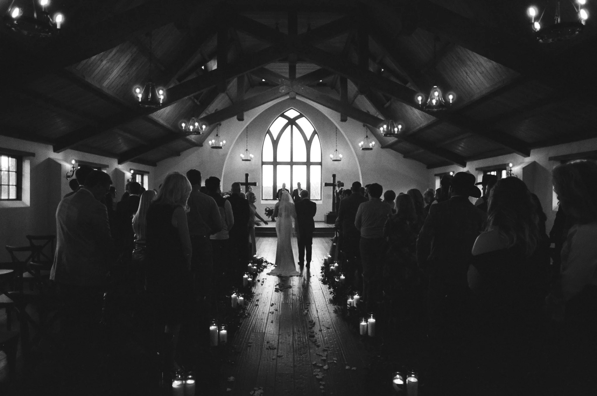 A black-and-white photo of a wedding ceremony inside a church with wooden architecture. The bride and groom stand at the altar, facing an arched window with sunlight streaming through. Guests are seated or standing on either side, and there are lit candles along the aisle.