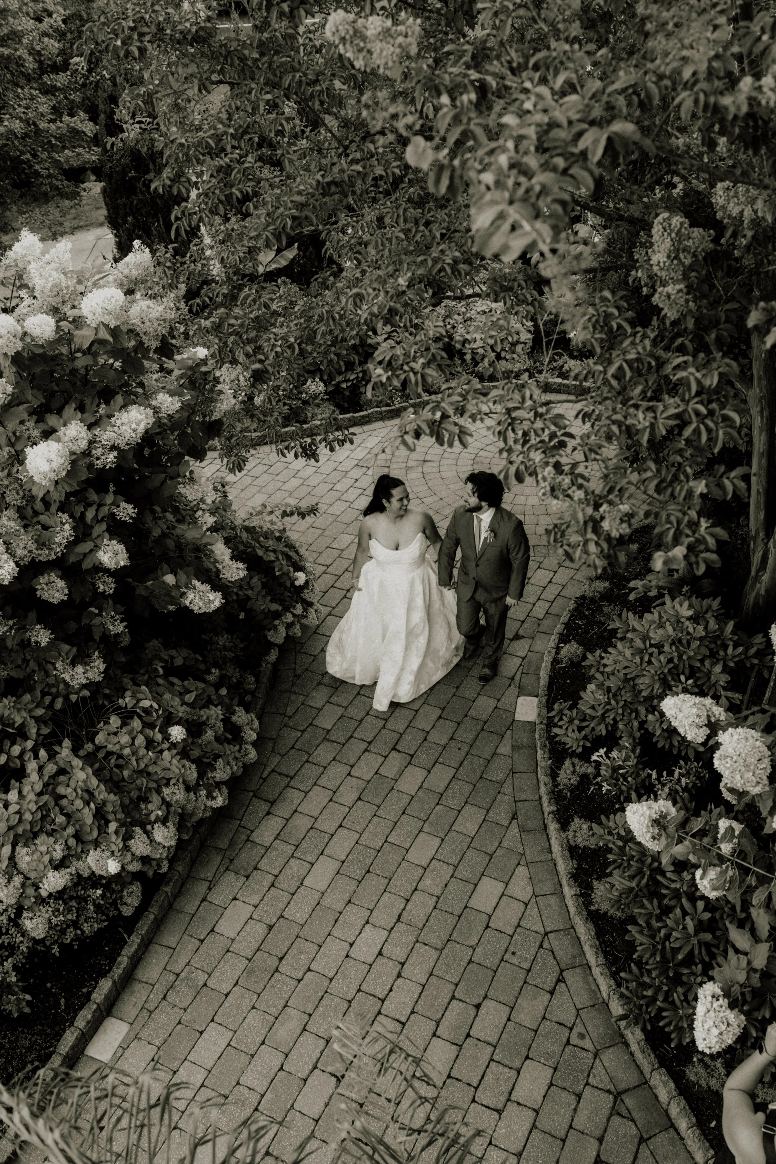 A black and white photo of a bride and groom walking hand in hand on a brick pathway surrounded by lush garden foliage and flowers.