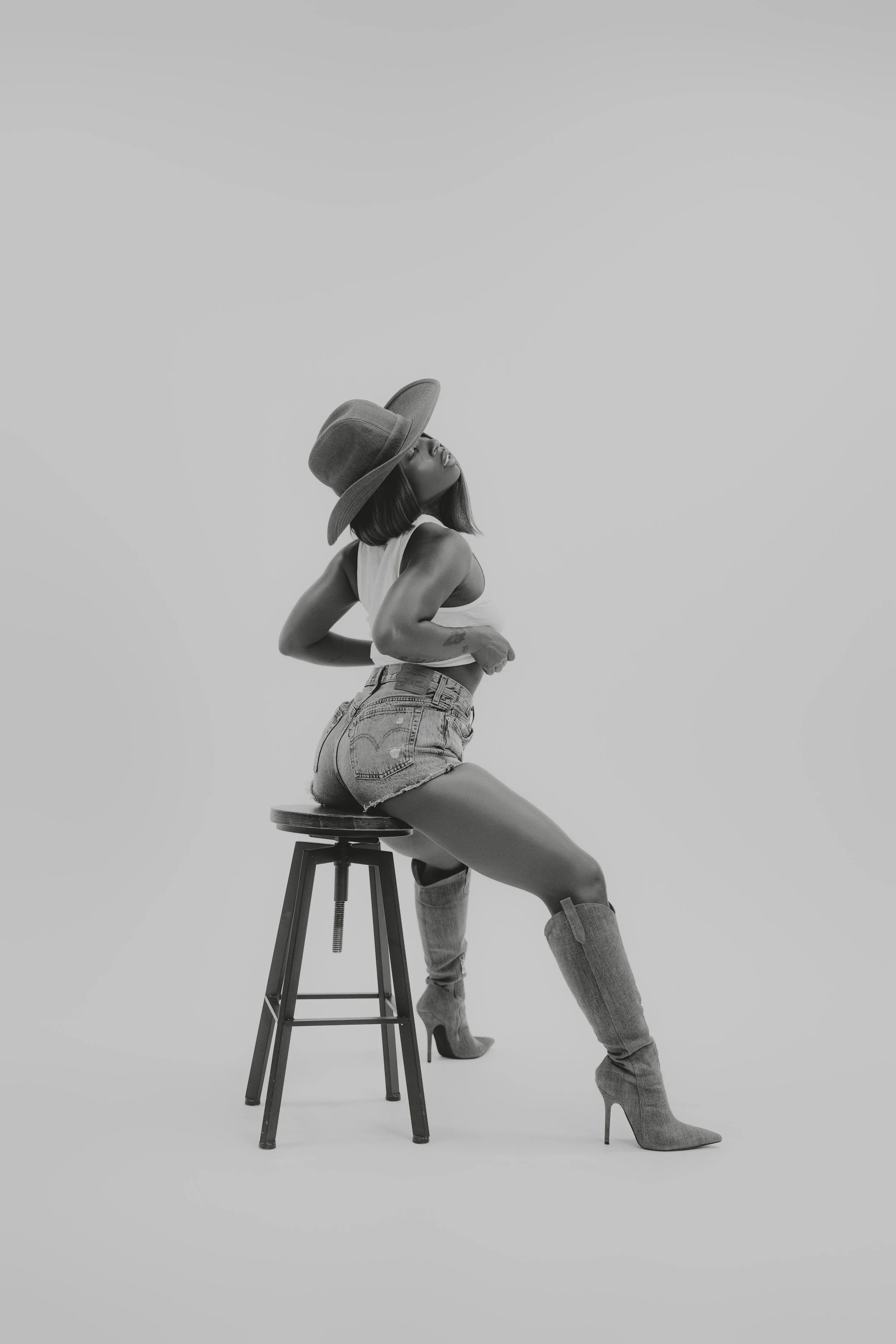 A woman wearing a wide-brimmed hat, sleeveless top, denim shorts, and high-heeled boots, sitting on a stool in a minimalist studio setting.
