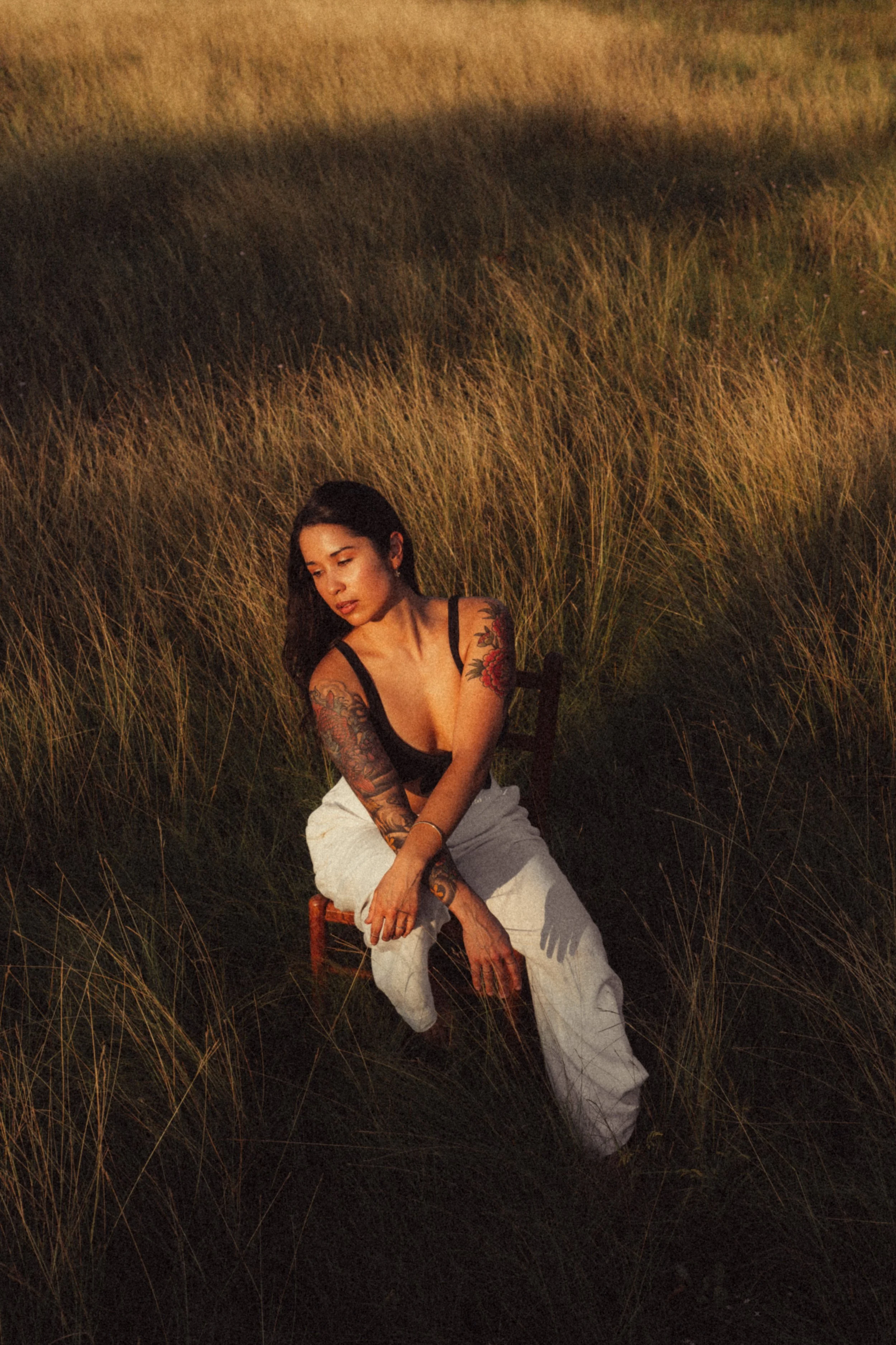 A woman with tattoos, wearing a black top and white pants, sitting on a chair in a field of tall grass during sunset.