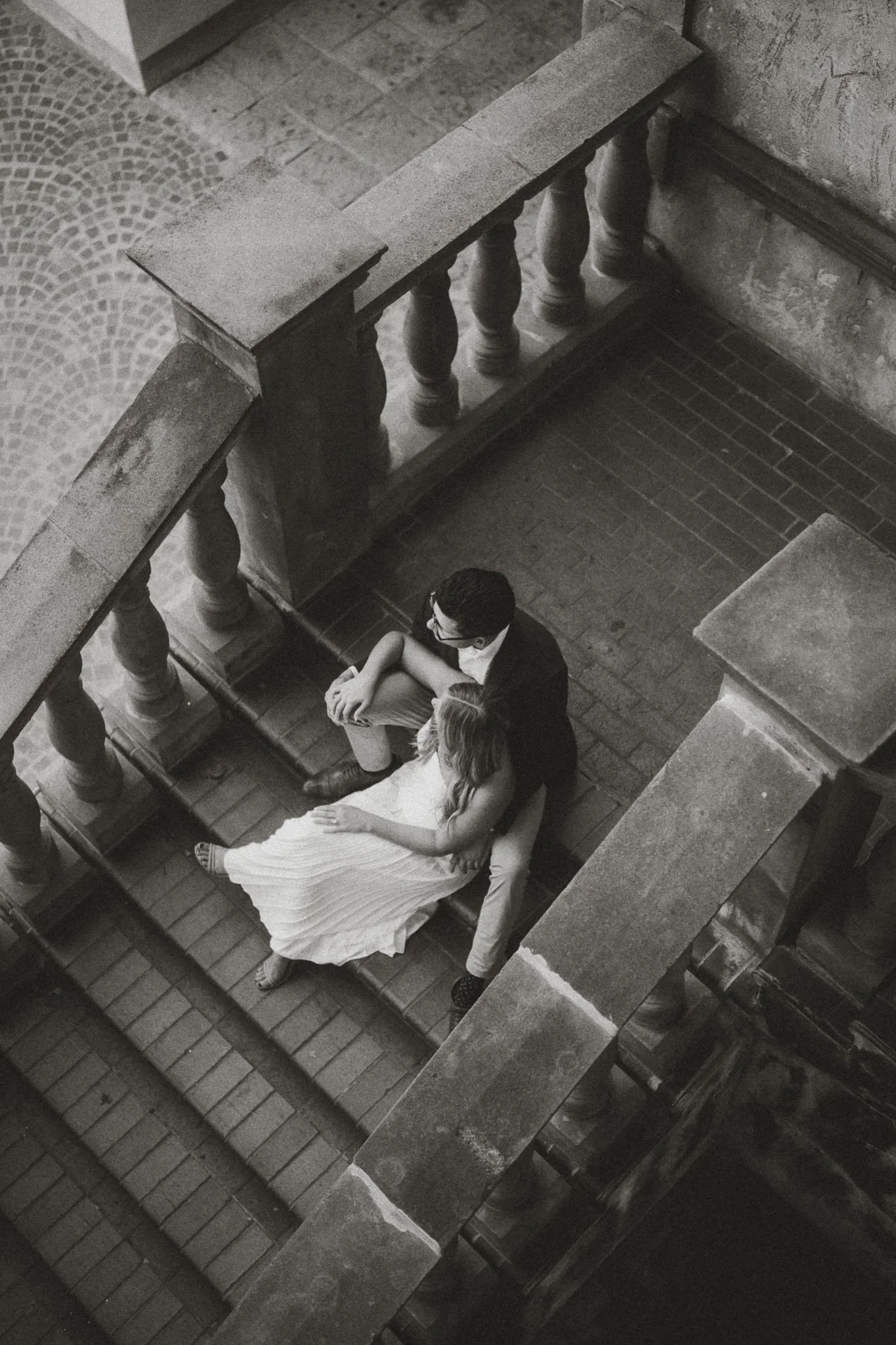 A black and white photo of a man and woman sitting on a staircase with stone balustrades in an indoor setting. The woman is lying back with her head on the man's lap, and they are looking at each other.