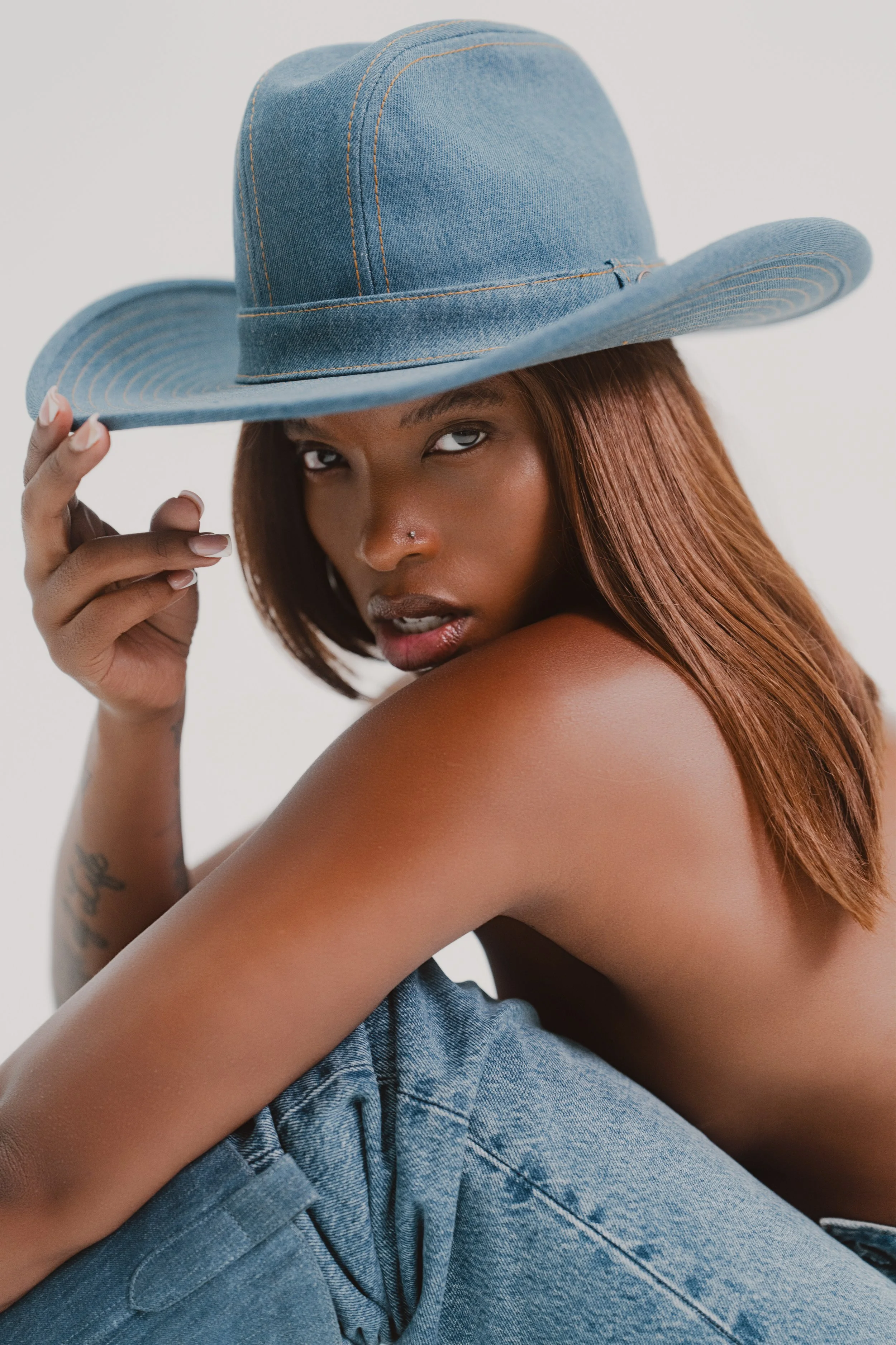 A woman with reddish-brown hair wearing a blue denim wide-brimmed hat, with her top off and sitting with one knee up, looking at the camera.
