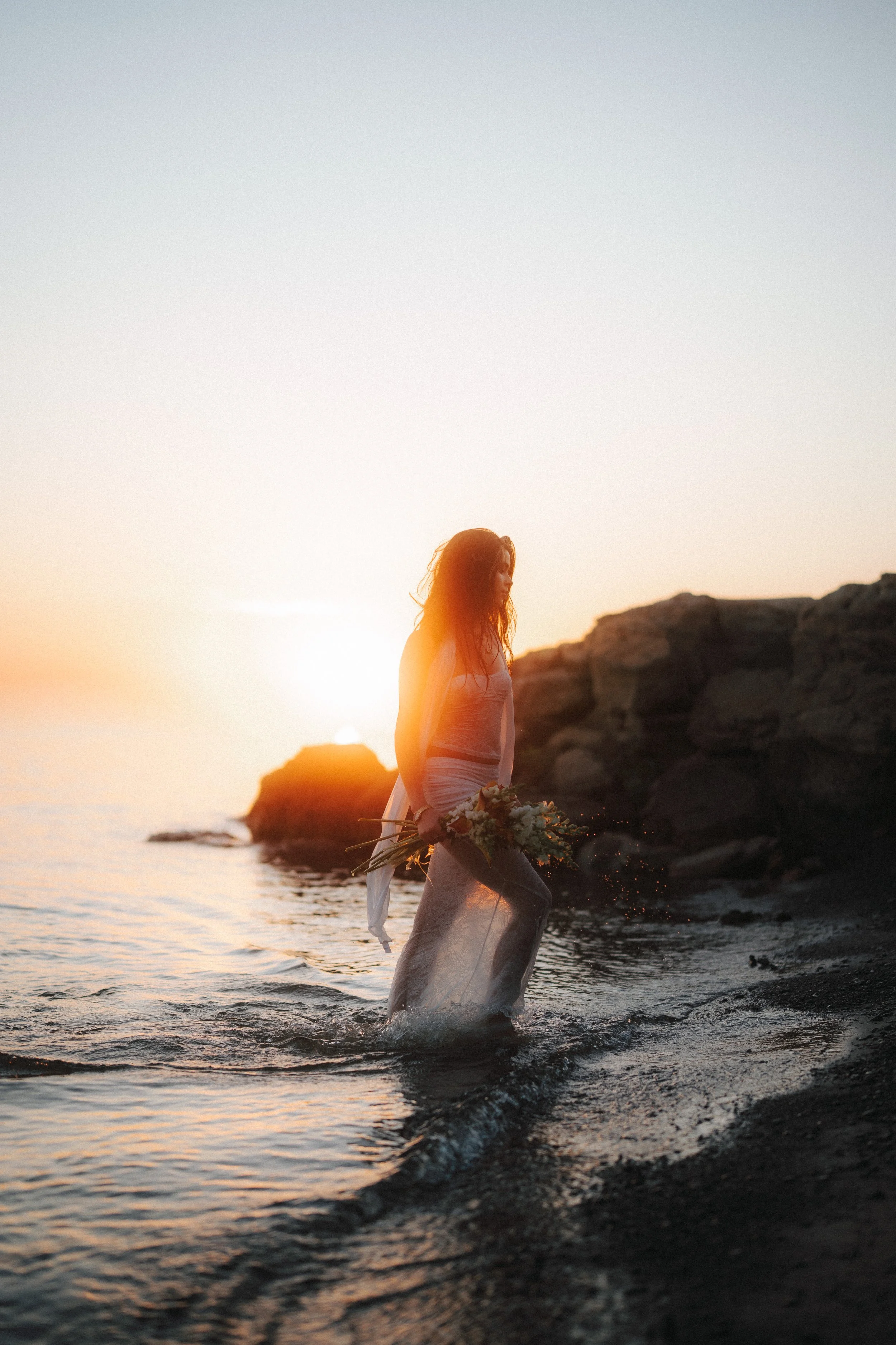 A woman standing in shallow water at the beach during sunset, holding a bouquet of flowers, with rocky formations in the background.