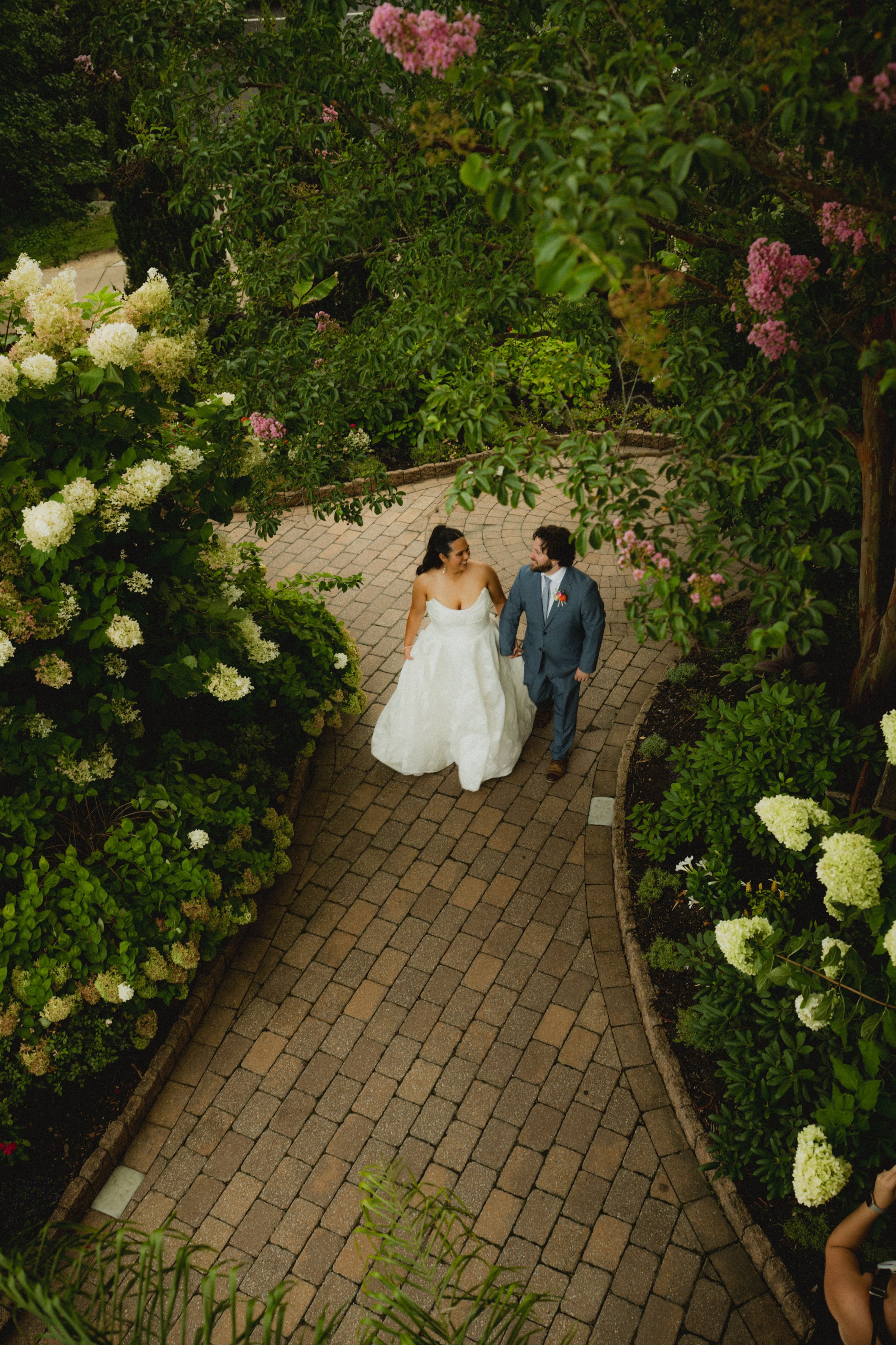 Bride and groom walking hand in hand on a brick pathway, surrounded by lush green bushes and blooming white and pink flowers, during a wedding celebration.