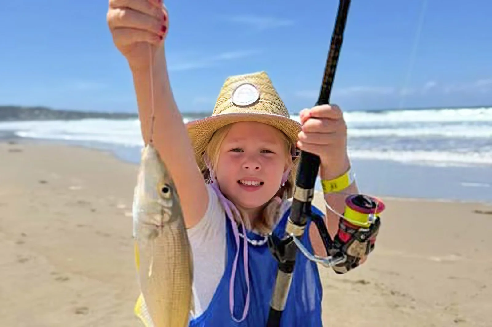 A smiling child on the beach proudly holding up a freshly caught fish while fishing with a local guide, with waves rolling in behind them