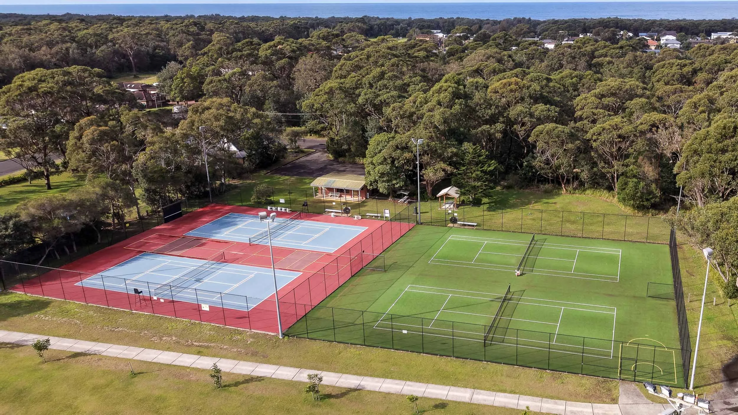 A wide-angle aerial shot of the Shoalhaven Heads tennis club facility, featuring multiple courts, a clubhouse building, and the surrounding trees and coastline in the distance.