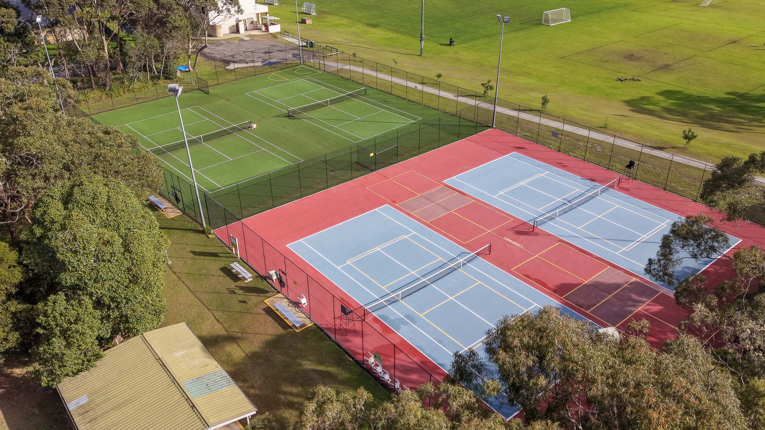 An aerial view of the Shoalhaven Heads tennis and pickleball courts, showing the blue and red court surfaces, fencing, and surrounding green fields.