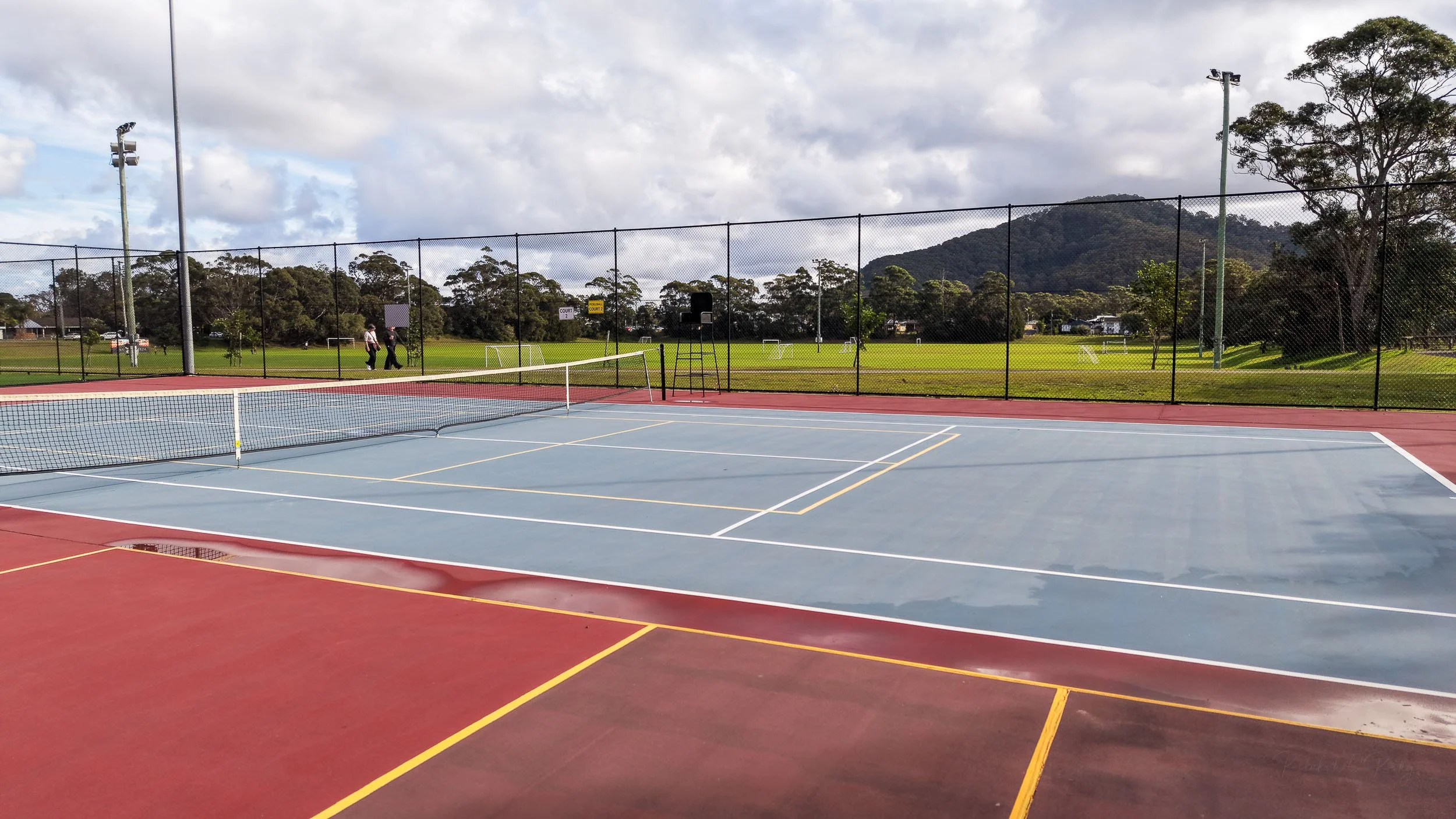 A ground-level view of a blue and red tennis court at the Shoalhaven Heads club, with a net in the foreground and parkland in the background.