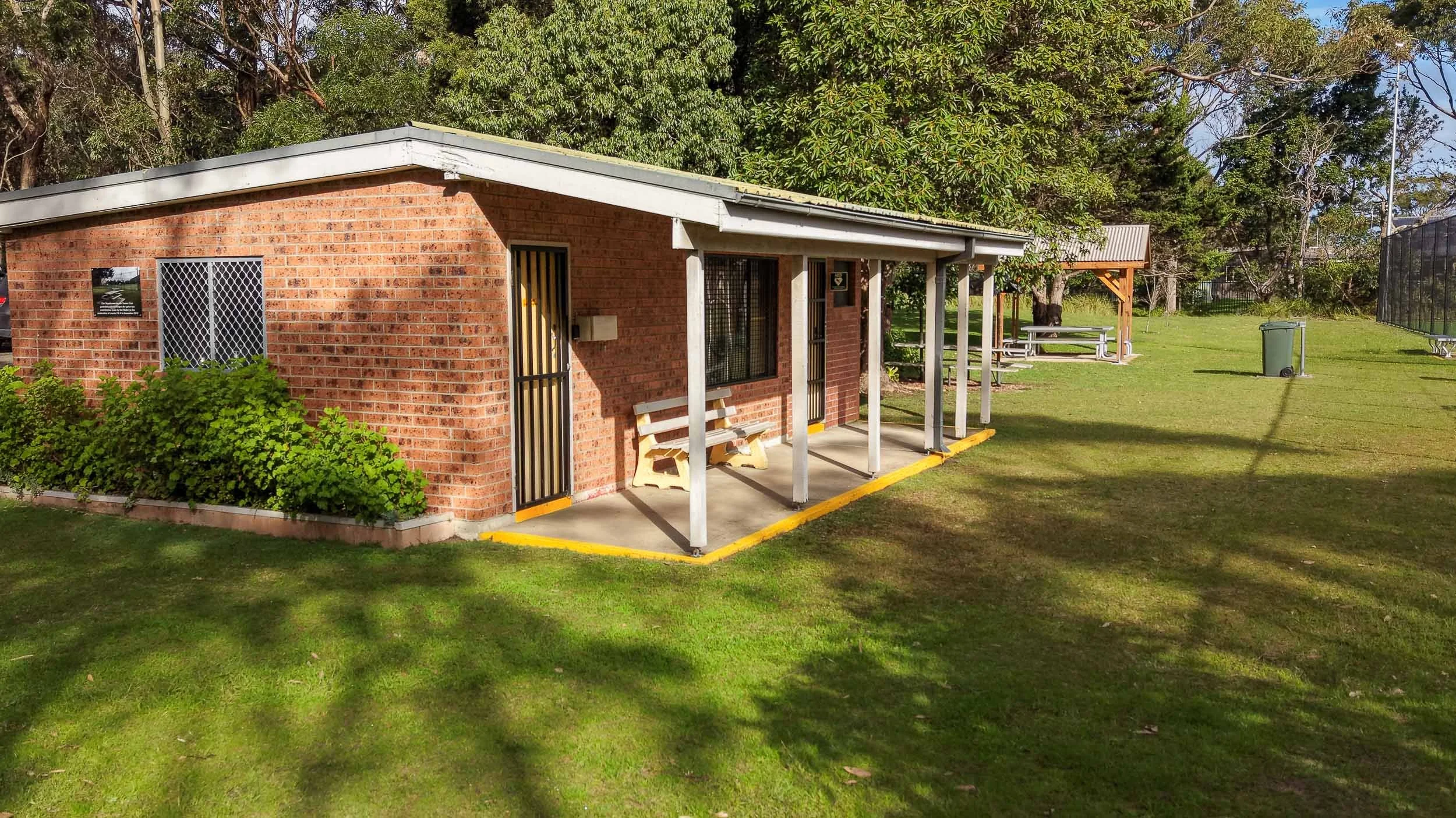 A close-up of the brick clubhouse building at the Shoalhaven Heads Tennis Club, showing its covered porch, benches, and surrounding grassy area.