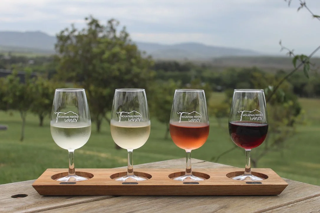 A flight of four wine glasses presented in a row on a long wooden paddle tasting board, resting on a rustic wooden table. The glasses contain a gradient of liquids from left to right: a pale translucent white, a light golden white, a vibrant pink ros