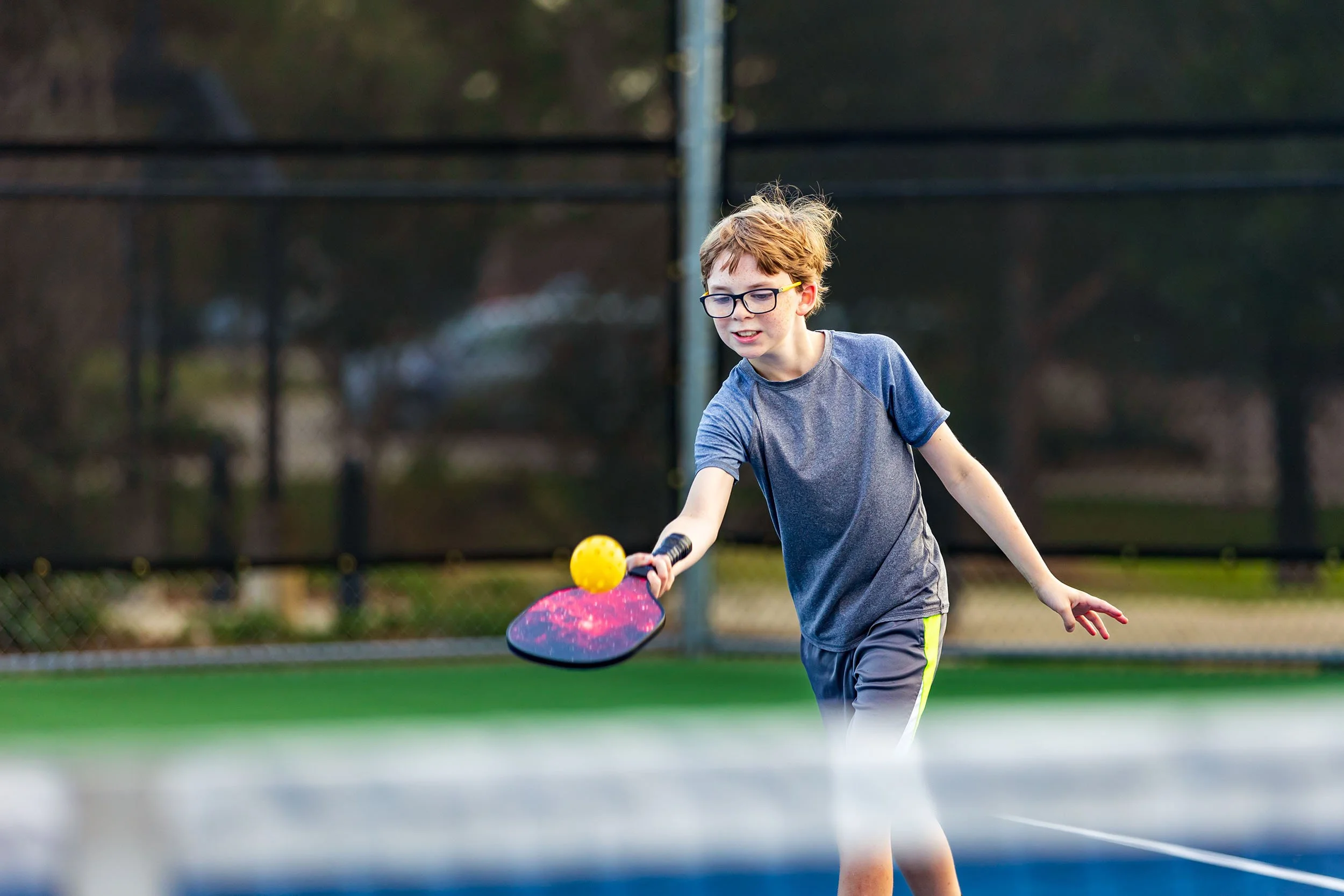 A young boy with glasses hits a yellow pickleball with a paddle on a green outdoor court.
