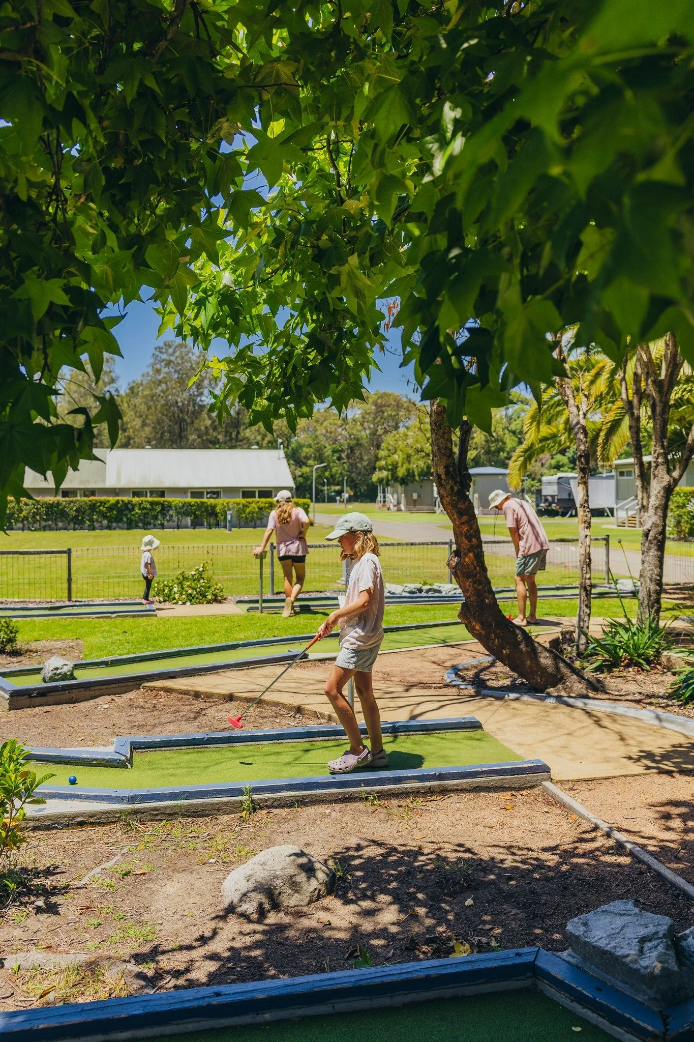 Children and adults playing miniature golf outdoors at Holiday Haven Shoalhaven Heads on a sunny day, surrounded by trees and green grass.