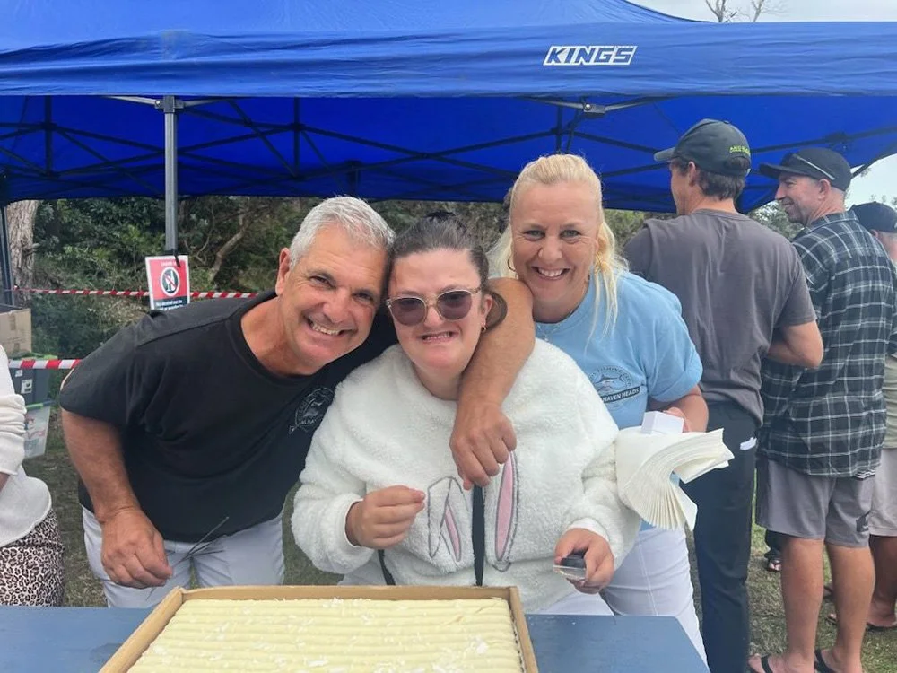 Three smiling club members posing behind a large cake at an outdoor event at Carters Corner.