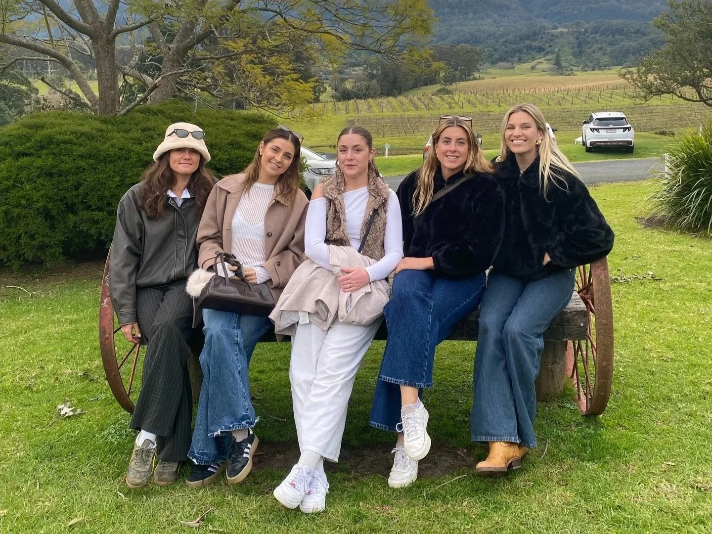 A group of five friends sitting together outdoors on a wooden wagon wheel with scenic vineyard rows and coastal hills in the background in the Shoalhaven region.