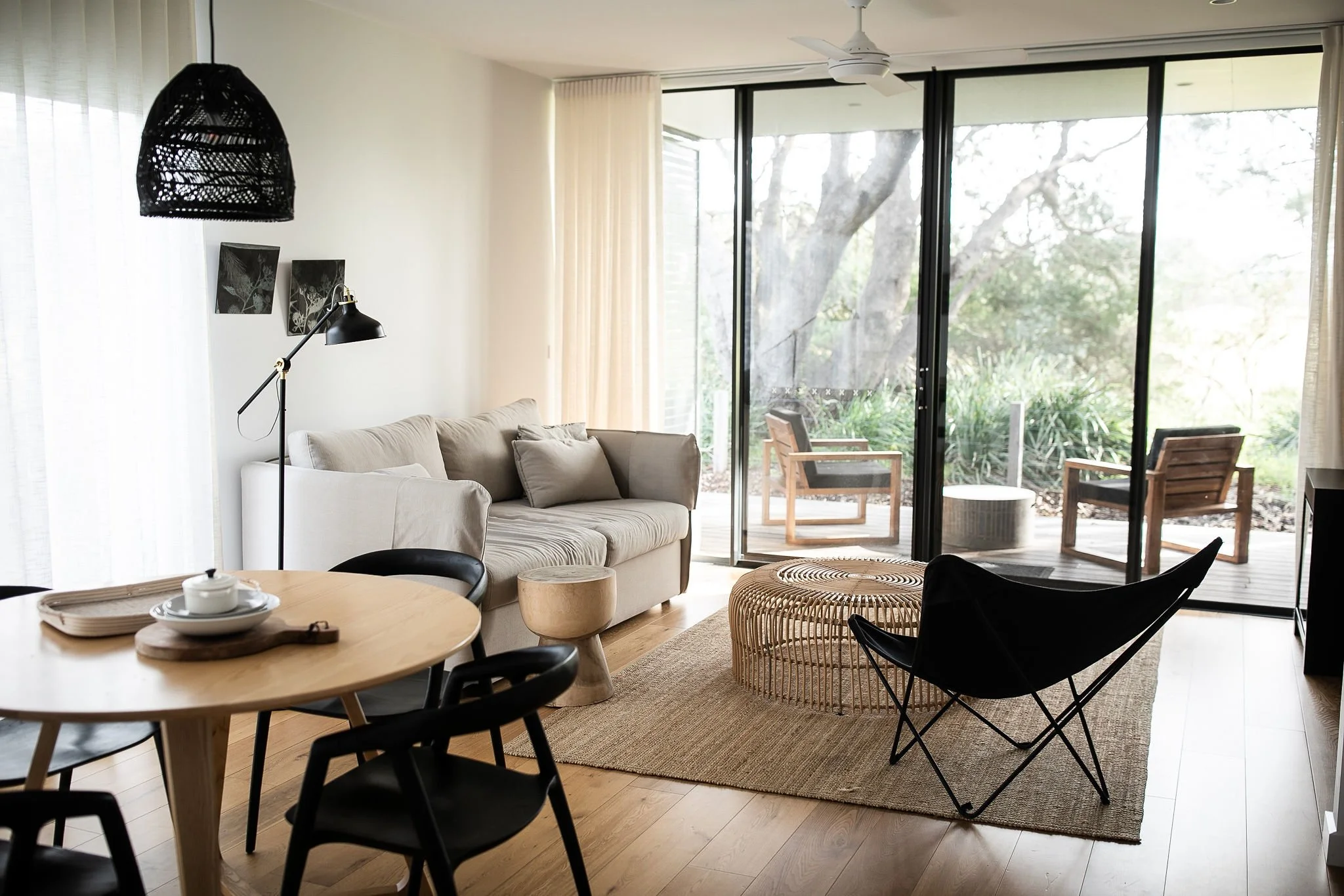 Modern living room with beige sofa, black armchair, round wicker coffee table, wooden side table, black floor lamp, black hanging pendant light, wall art, and sliding glass door leading to a patio with outdoor chairs and greenery at Bangalay Villas.