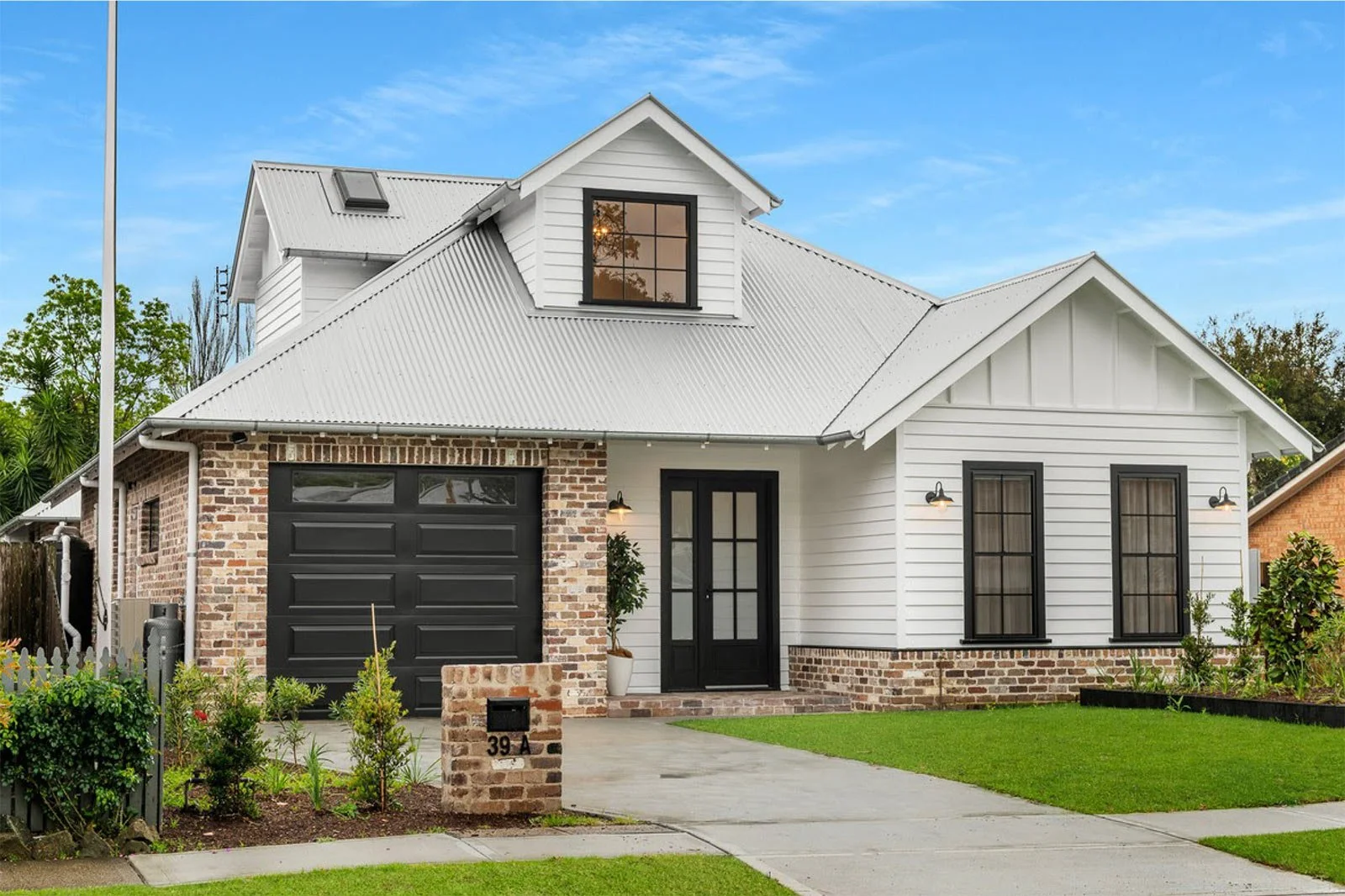 White weatherboard cottage-style home with a grey corrugated roof, black-framed windows, and exposed brick accents, featuring a black garage door and landscaped front yard.
