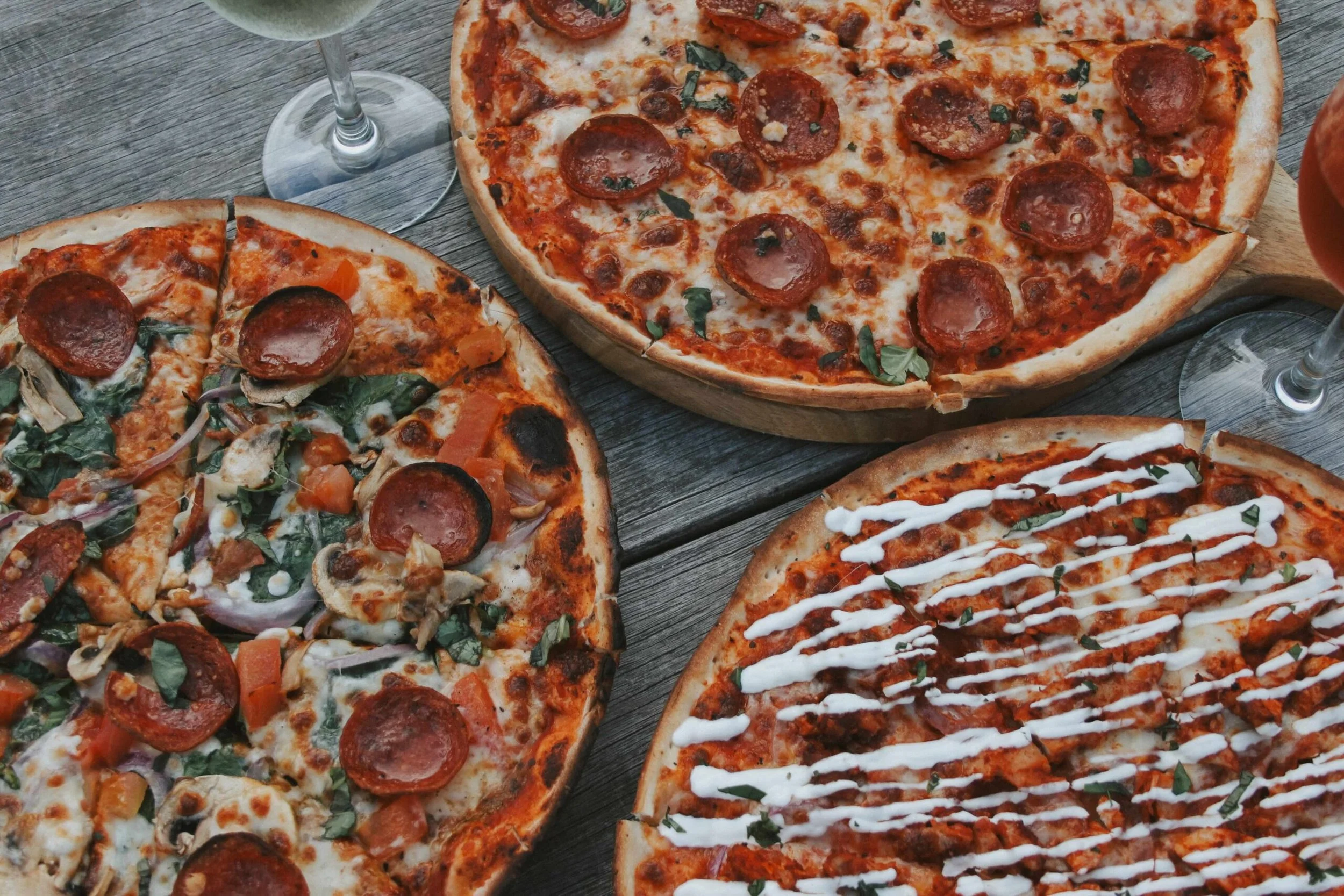 A top-down, close-up shot of three large, rustic, thin-crust pizzas arranged on a weathered wooden table. The pizza on the top is topped with pepperoni and fresh basil; the pizza on the left is loaded with pepperoni, mushrooms, red onion, spinach, an
