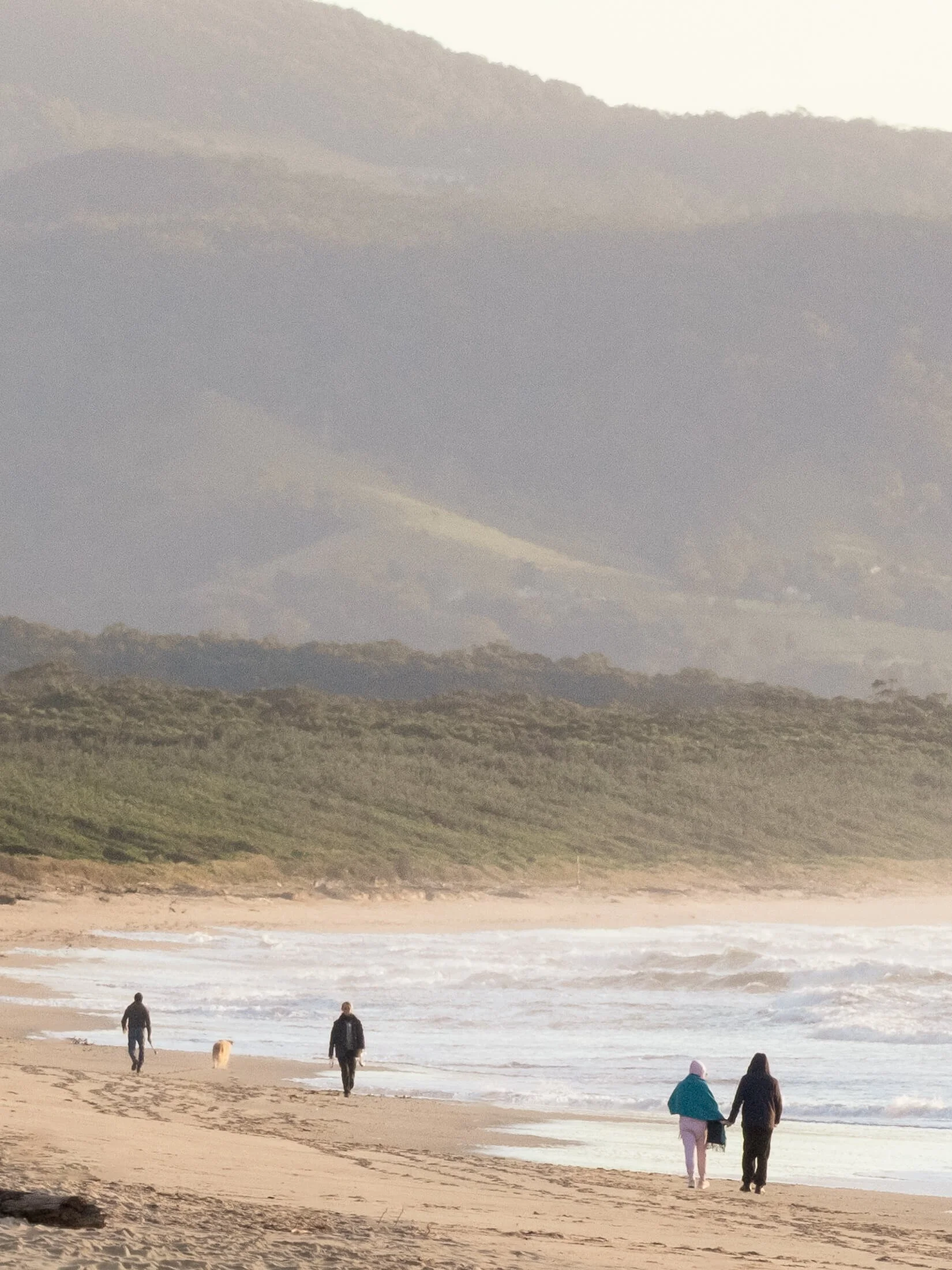 People walking along a Seven Mile Beach beach in Shoalhaven Heads with ocean waves and green hills in the background during daylight.