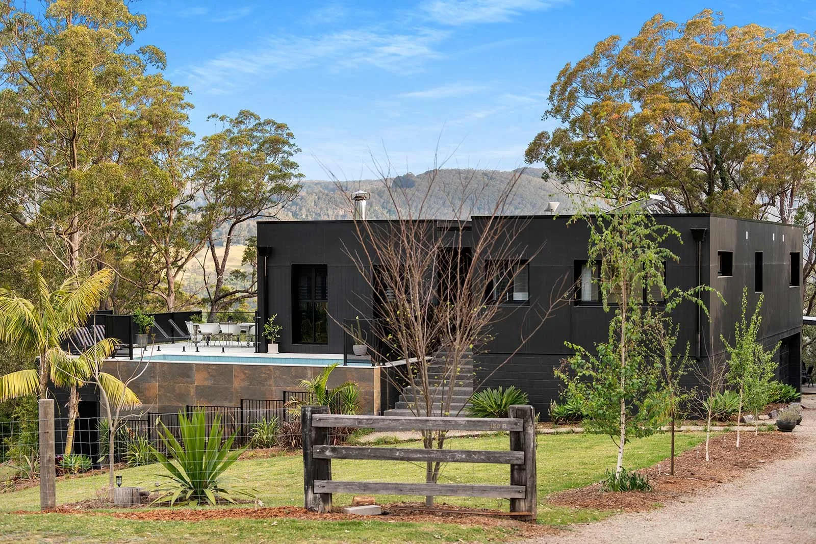 Modern black rectangular home with large windows, surrounded by mature gum trees and landscaped gardens, with a pool and outdoor seating visible behind a rustic wooden gate.