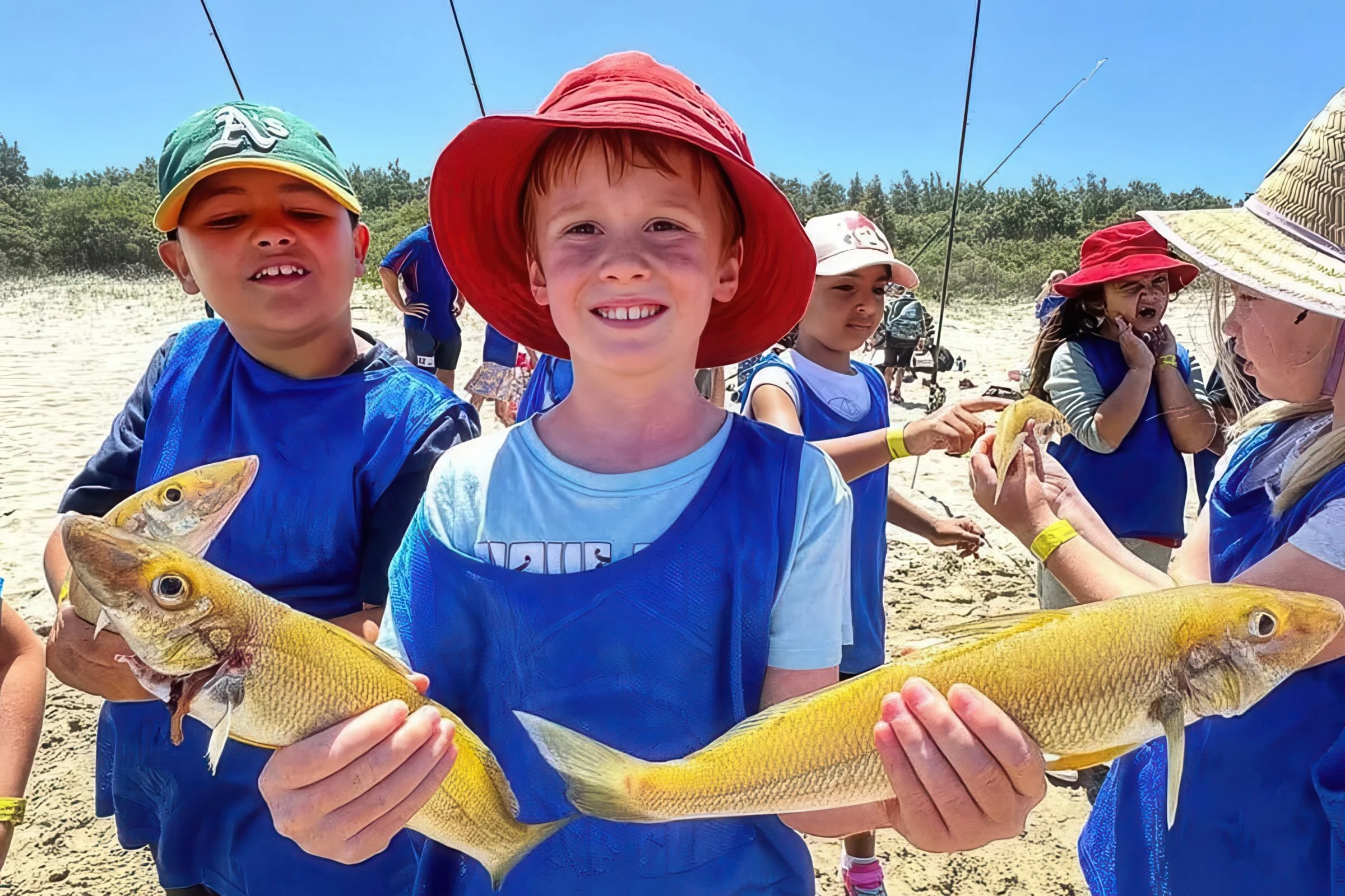 Kids on a sunny beach wearing blue fishing vests, smiling and proudly holding freshly caught fish during a Fishing With A Local lesson.