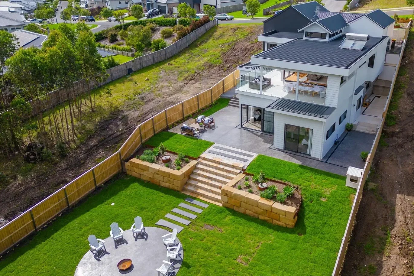 Modern multi-level white home with black roof accents, overlooking a landscaped tiered backyard featuring stone retaining walls, wide steps, a circular firepit area and fresh green lawn.