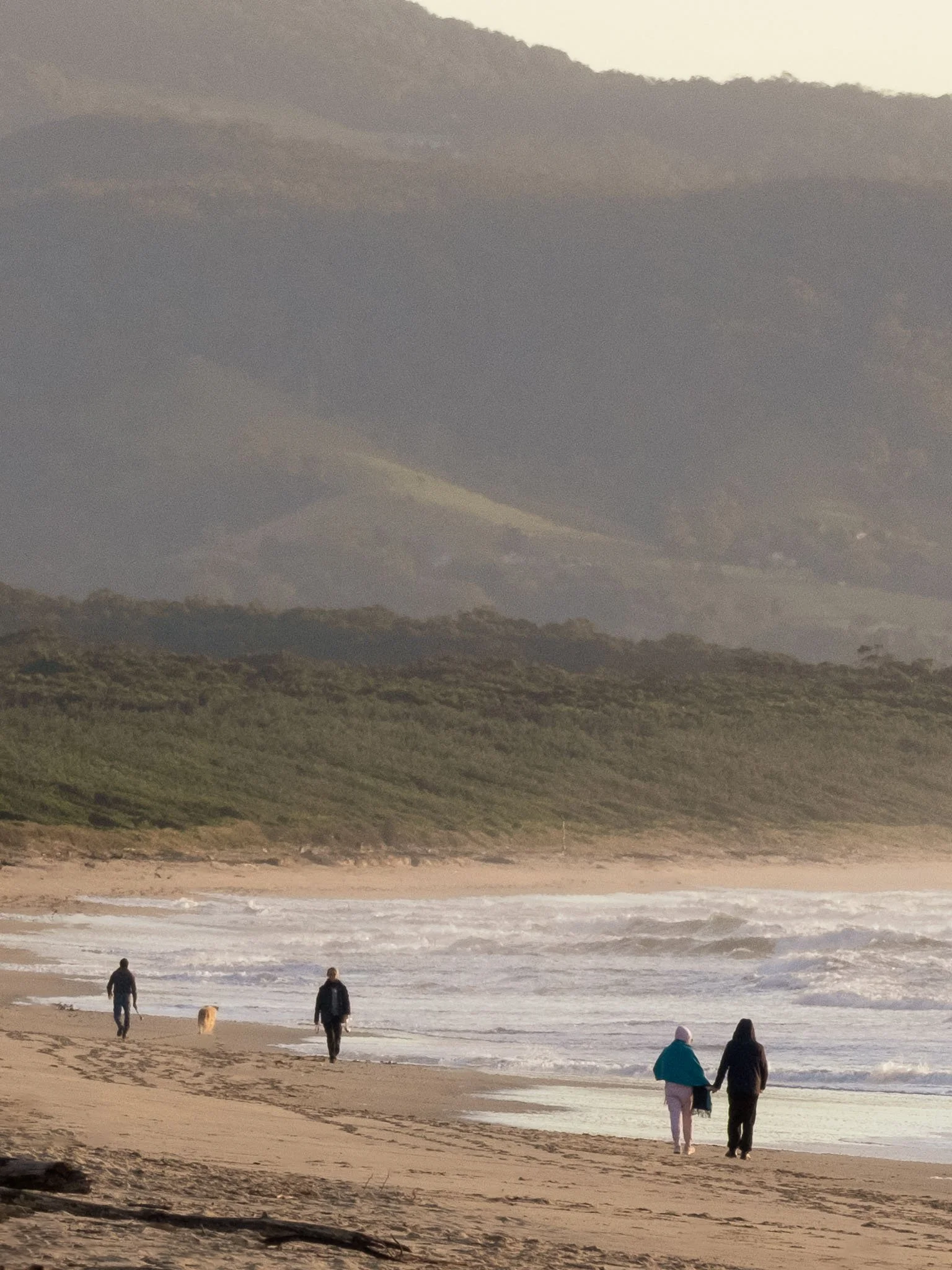 People walking along Seven Mile Beach at Shoalhaven Heads near the shoreline with waves, dunes, and mountains in the background.