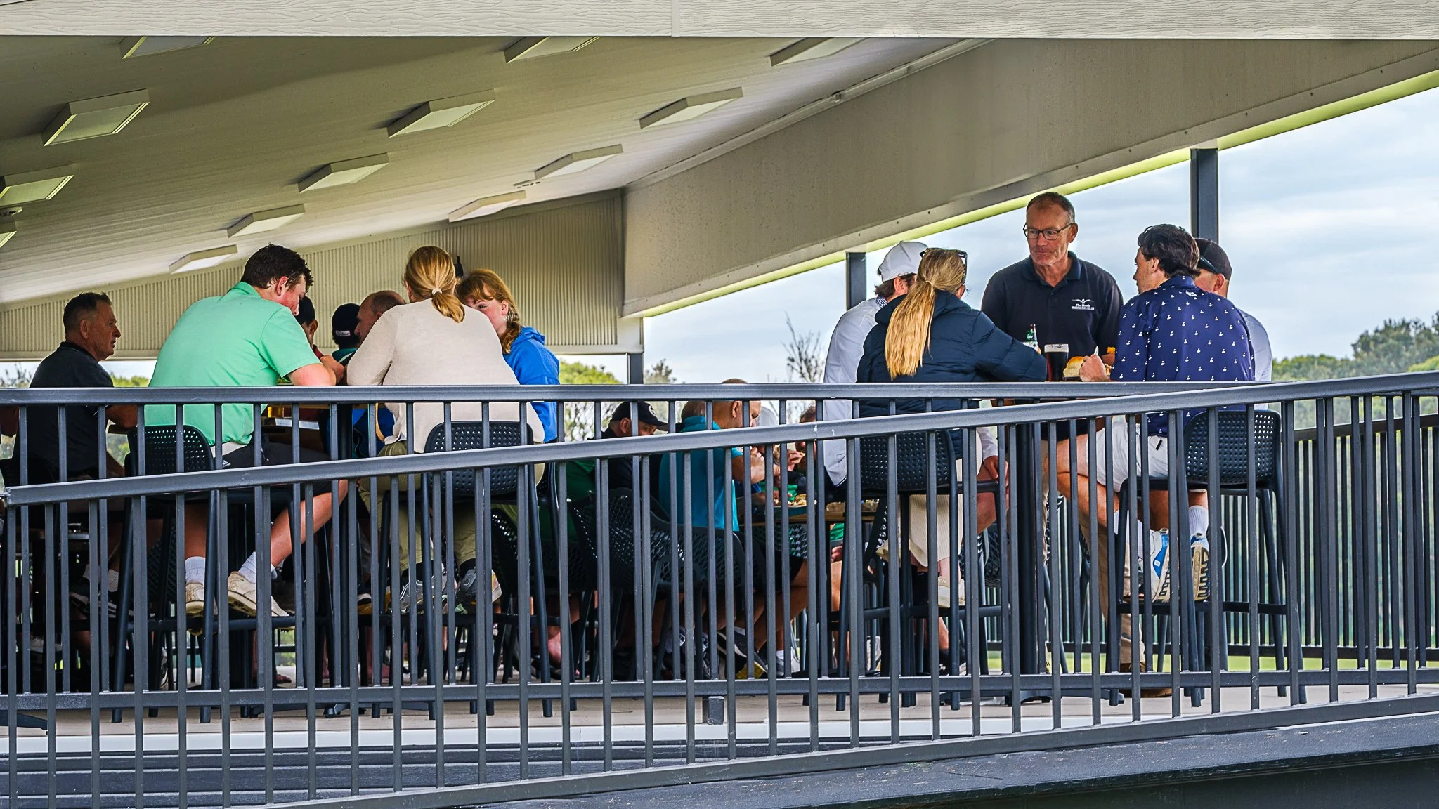 Group of people sitting on the verandah at Shoalhaven Heads Golf Club, with railing in front and trees in background.