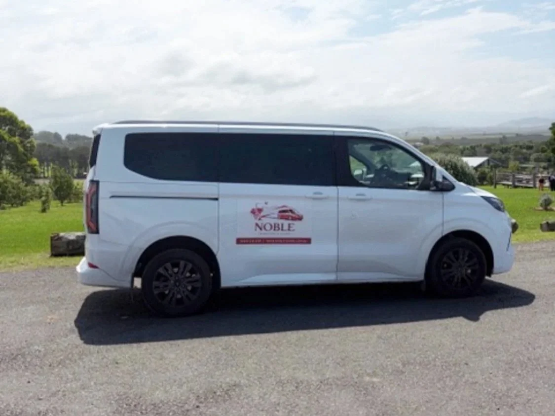 Side view of the official white Noble Transfers and Tours passenger van parked along a scenic rural roadside in the Shoalhaven region.