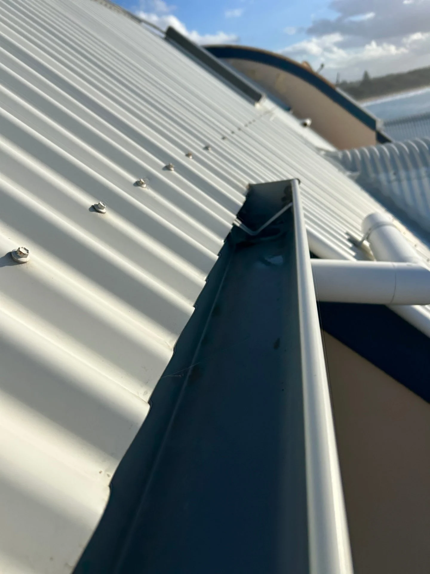 Close-up view of a freshly cleaned, empty Colorbond gutter installed on a white corrugated metal roof, with the coastline visible in the background.