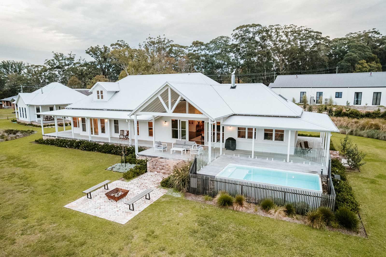 Large white country-style farmhouse with wraparound verandah, exposed gables and a small swimming pool, overlooking a spacious lawn with a brick firepit area and surrounding gardens.