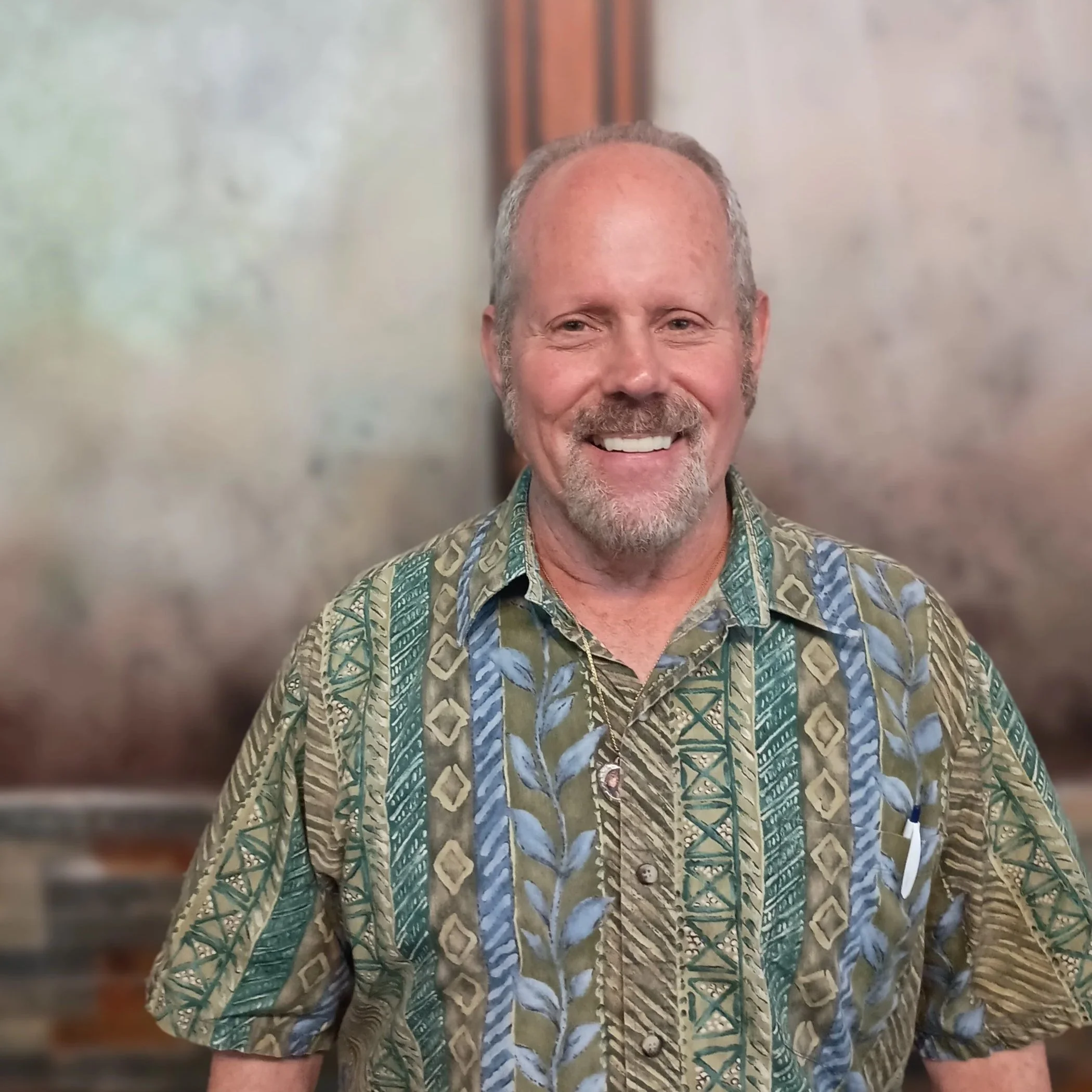 A smiling middle-aged man with a beard and mustache wearing a colorful patterned Hawaiian shirt.