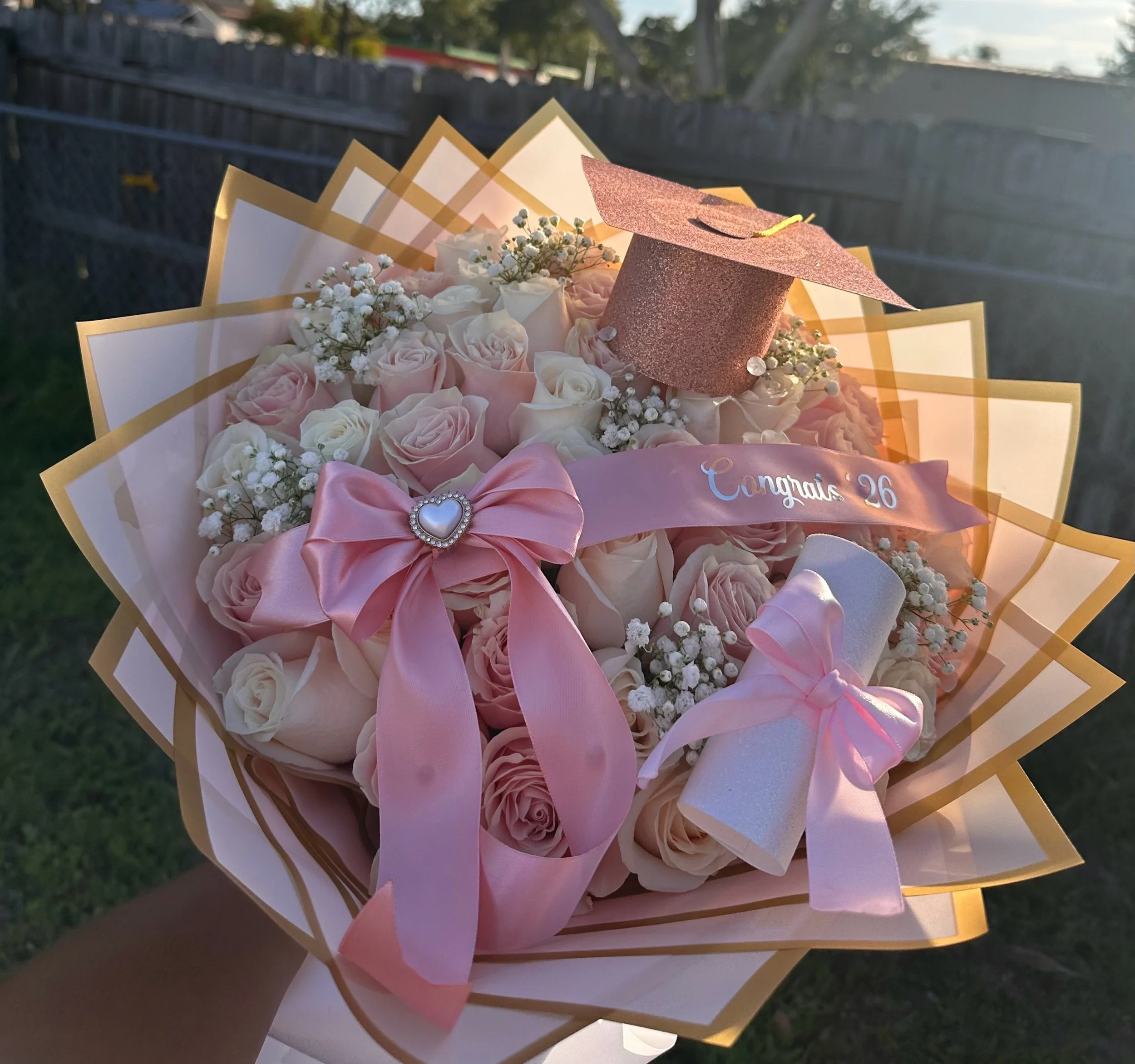 Pink and white rose graduation bouquet with baby’s breath, message banner, pink graduation cap, diploma roll, and pink wrapping with a gold border.