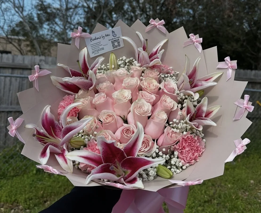 Pink rose bouquet with lilies, pink carnations, baby’s breath clusters, and mini pink bows, wrapped in brown floral paper.