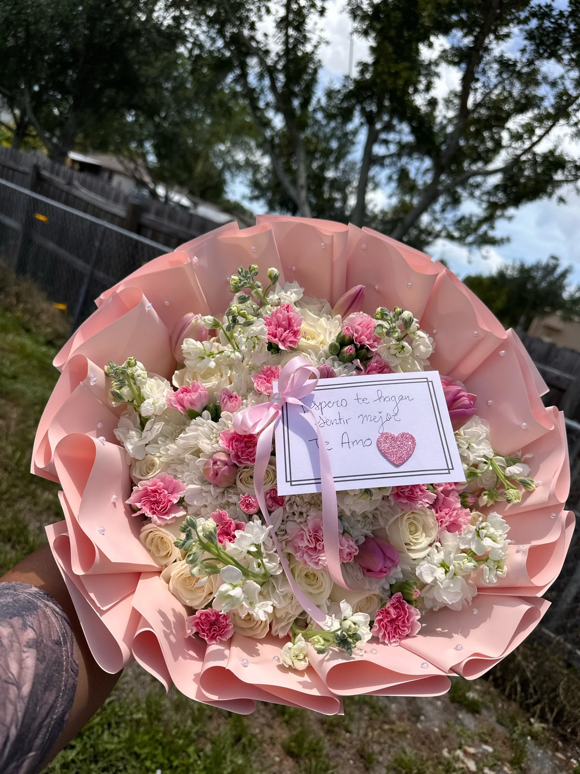 White rose bouquet with mini carnations, stock, poms, and tulips, wrapped in pink floral paper with pearl accents.