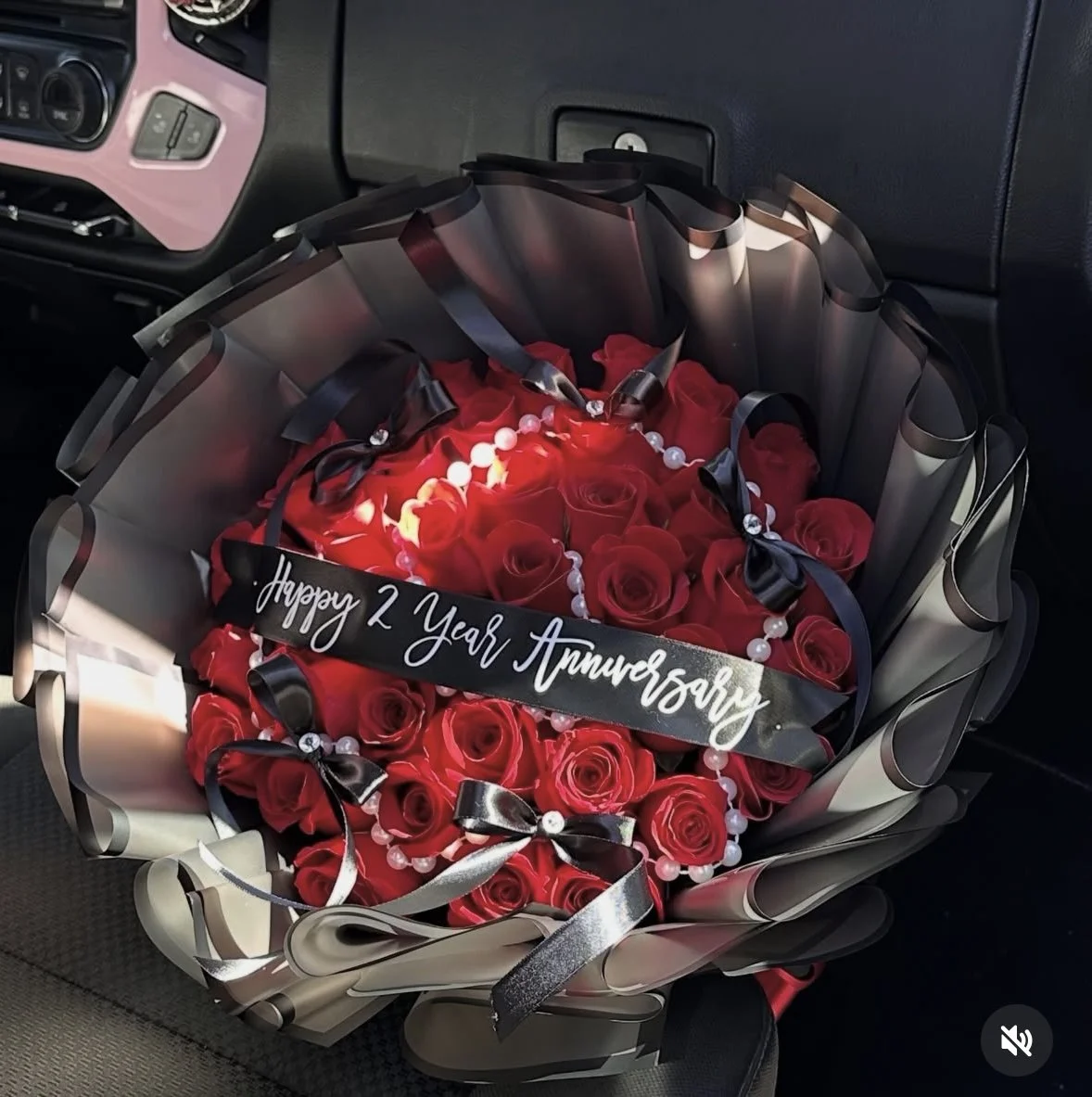 Red rose bouquet with pearl strands, long black bows, a message banner, and transparent black floral wrapping.
