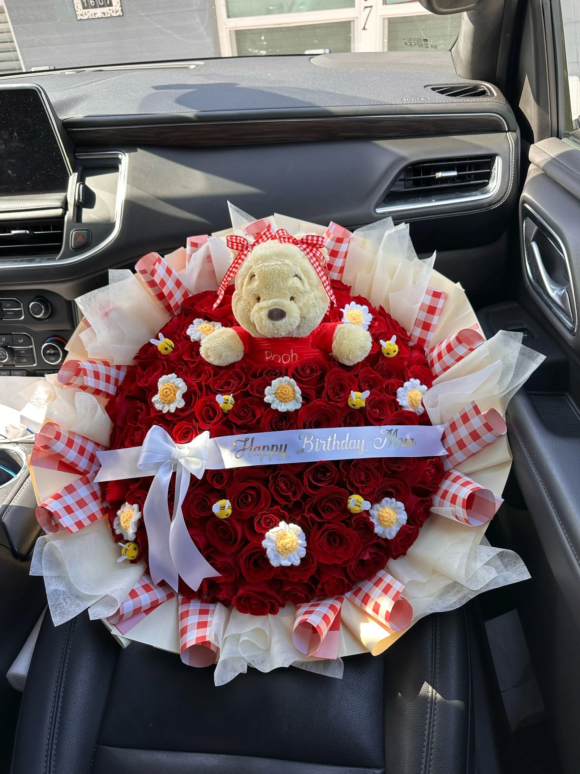 Red rose bouquet with crochet daisies, decorative bees, Winnie the Pooh plush, and yellow-and-red gingham floral wrapping.