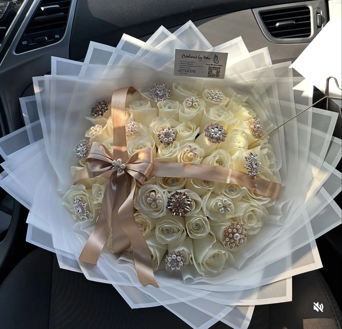 White rose bouquet with diamond pins, rhinestone accents, and a brown ribbon cross, wrapped in purple and white transparent floral wrapping.