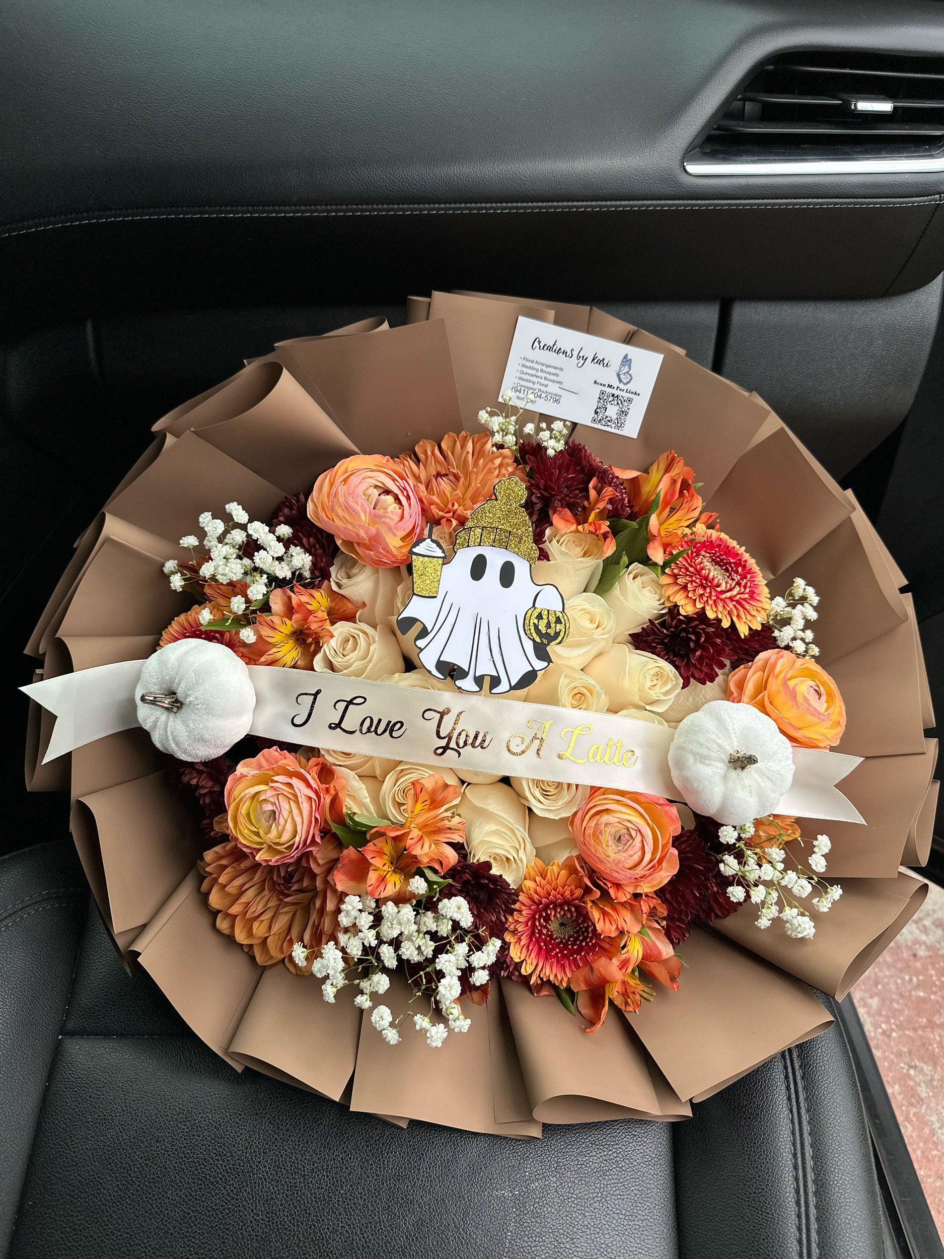 Fall bouquet with white roses, ranunculus, red poms, cremons, gerberas, baby’s breath, ghost topper, and pumpkins, wrapped in brown floral paper.