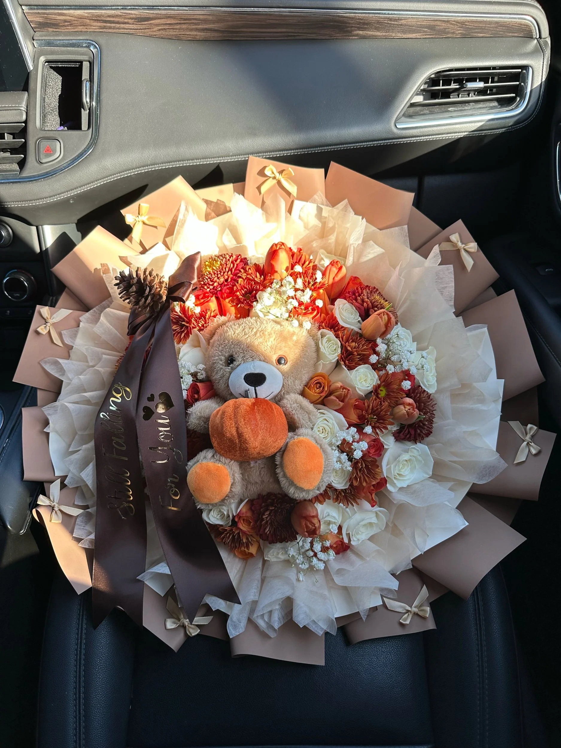 Orange and white rose bouquet with tulips, cremons, daisies, baby’s breath, a message bow, and teddy bear, wrapped in brown floral paper.