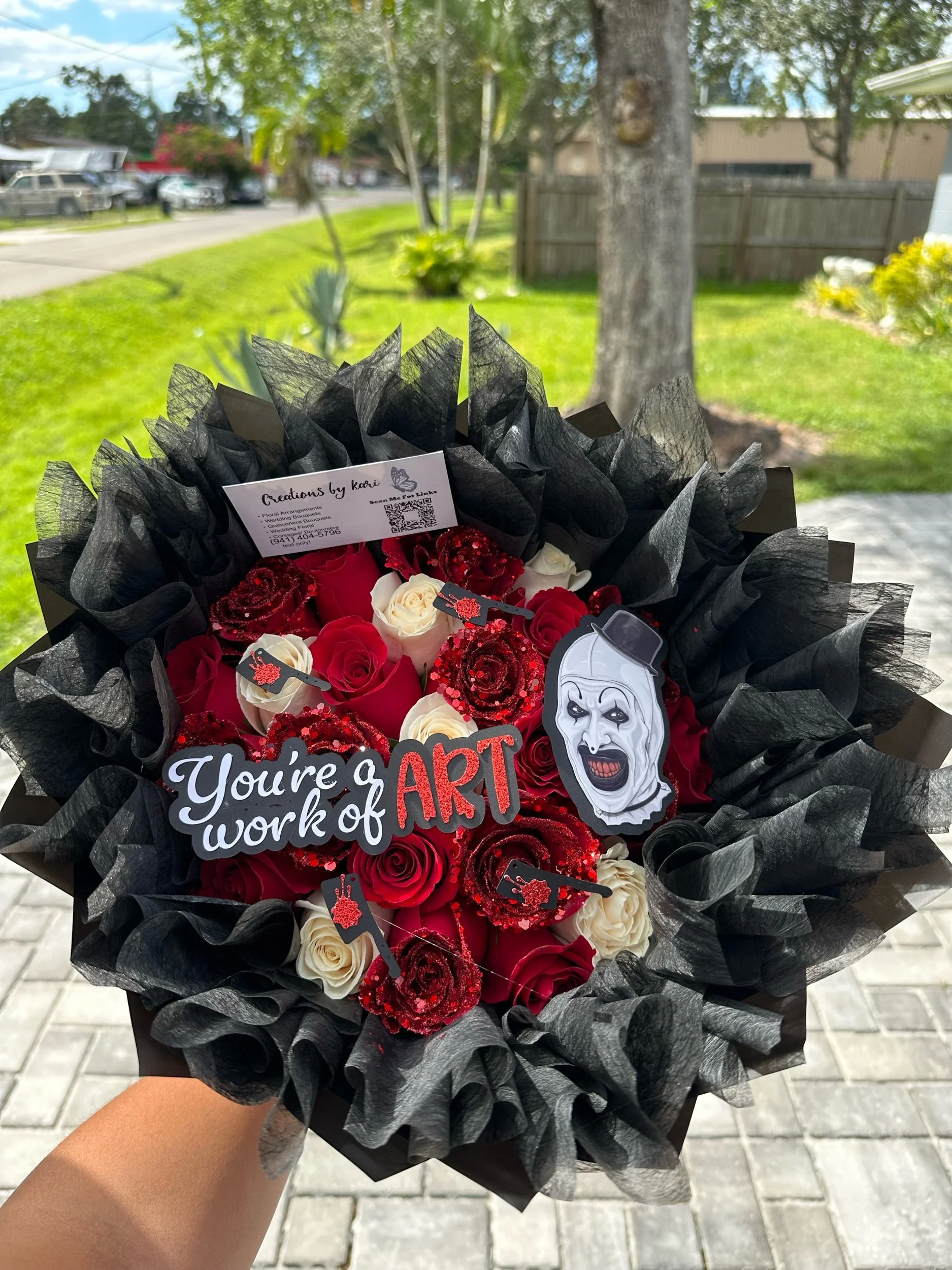 Red and white rose bouquet with glittered red roses, “You’re a Work of Art” message topper, Art the Clown topper, and black floral wrappin