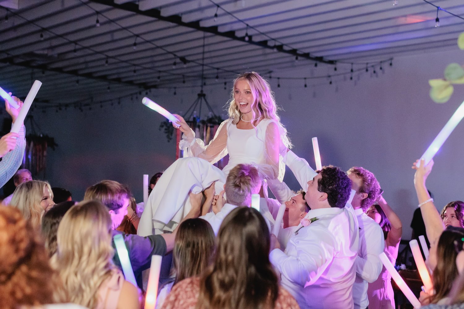 A joyful bride is lifted above the crowd during a high-energy wedding reception, surrounded by glowing light sticks and dancing guests. This celebration at our Utah wedding venue captures the fun, unforgettable moments that make your big day truly yo