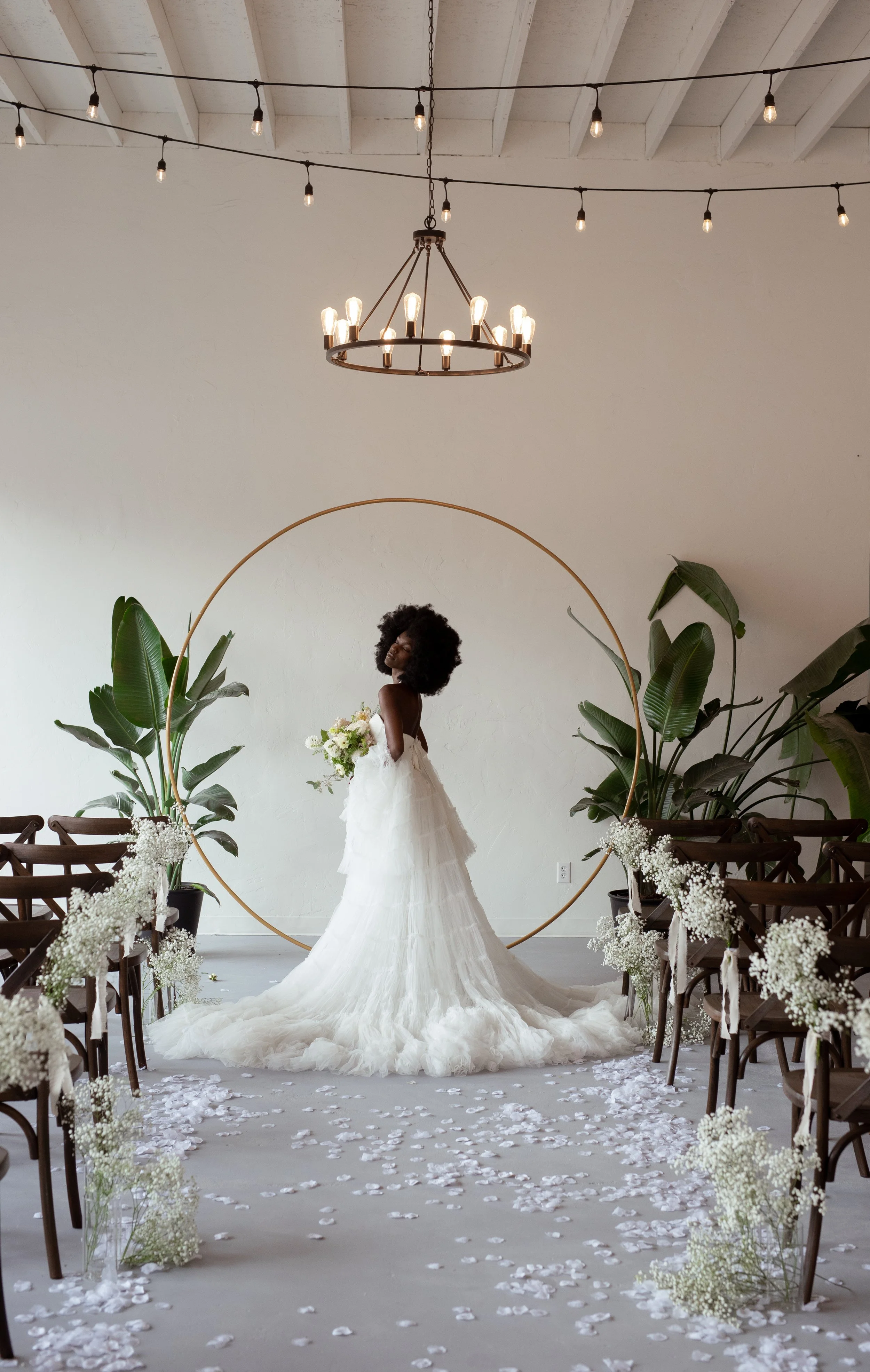 Elegant bridal portrait at ceremony arch

A bride in a flowing white gown stands beneath a gold circular arch adorned with greenery and florals, framed by soft white curtains.