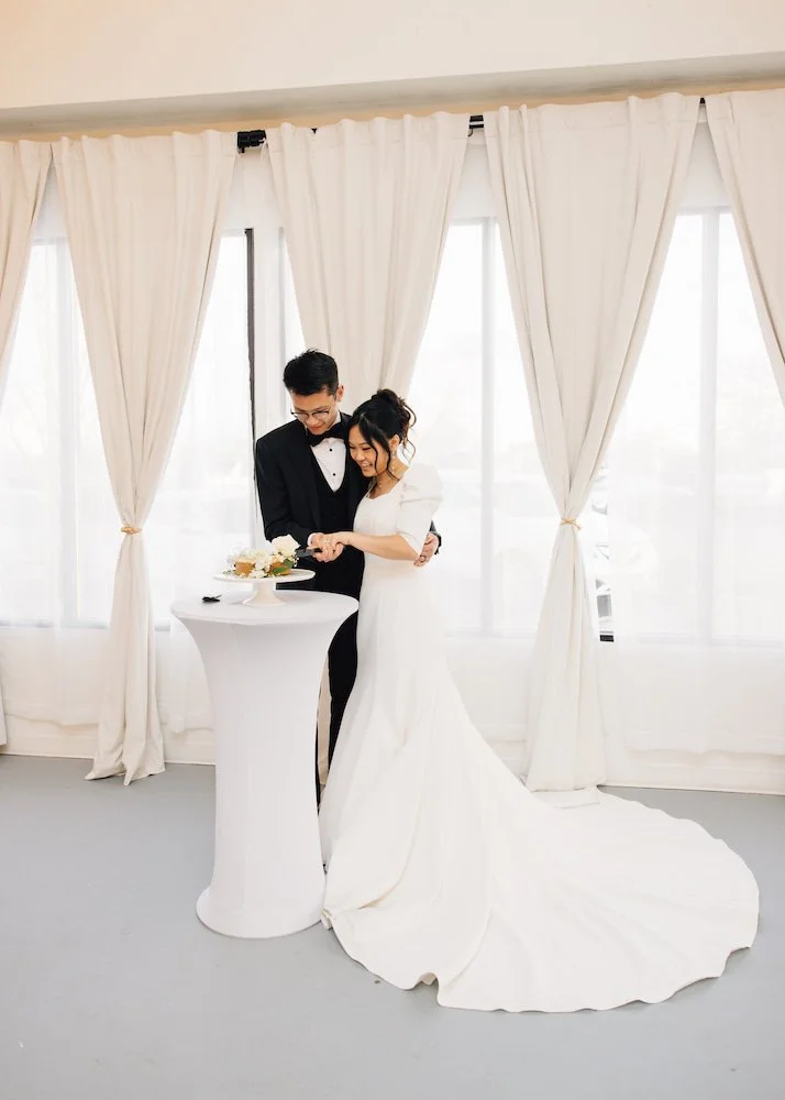 Bride and groom laugh together while cutting their wedding cake during their reception at The Ivory Hall, framed by beautiful soft-drape curtains.