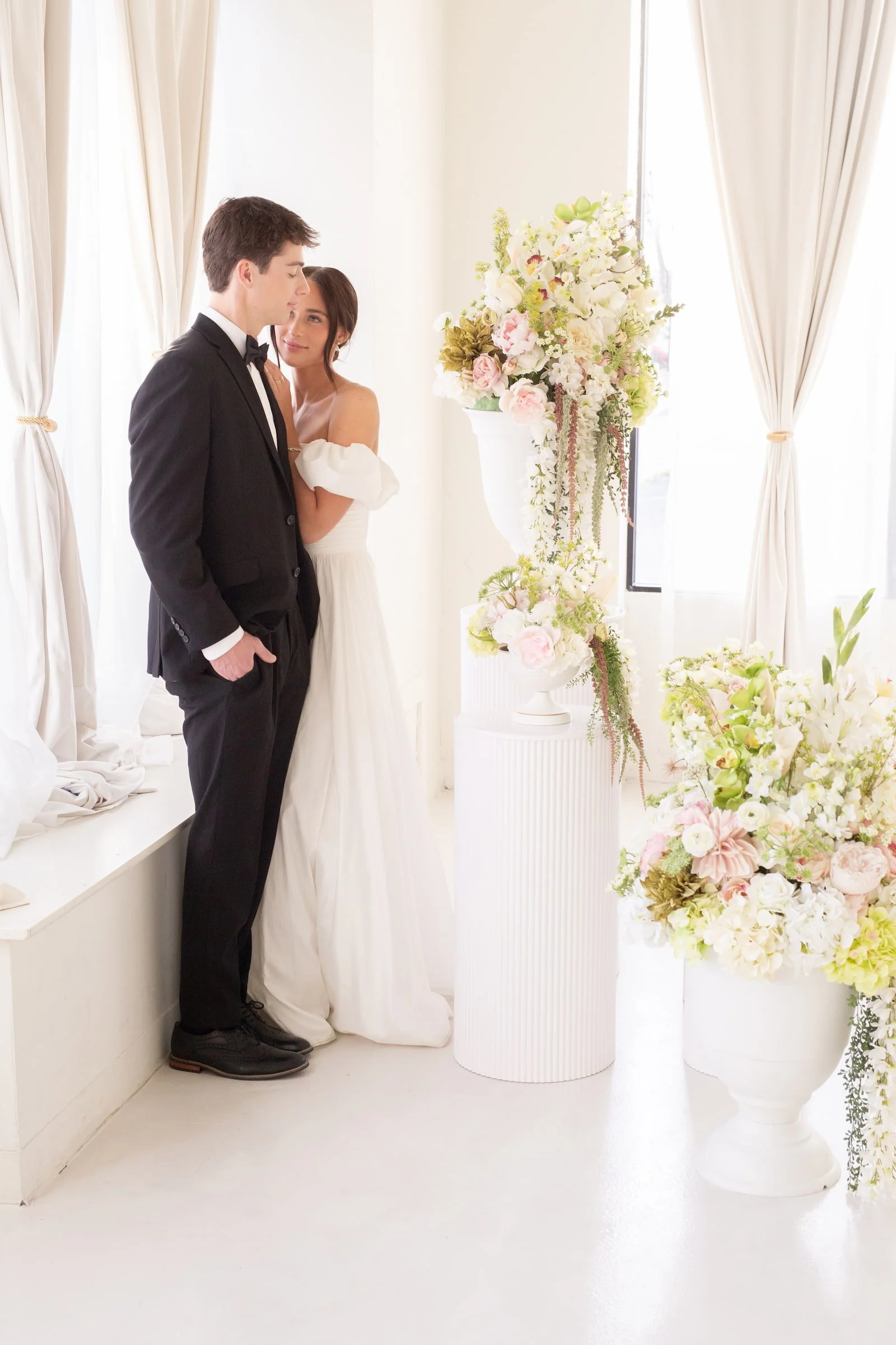 Bride and groom standing by tall windows with floral arrangements at The Ivory Hall in Provo, Utah