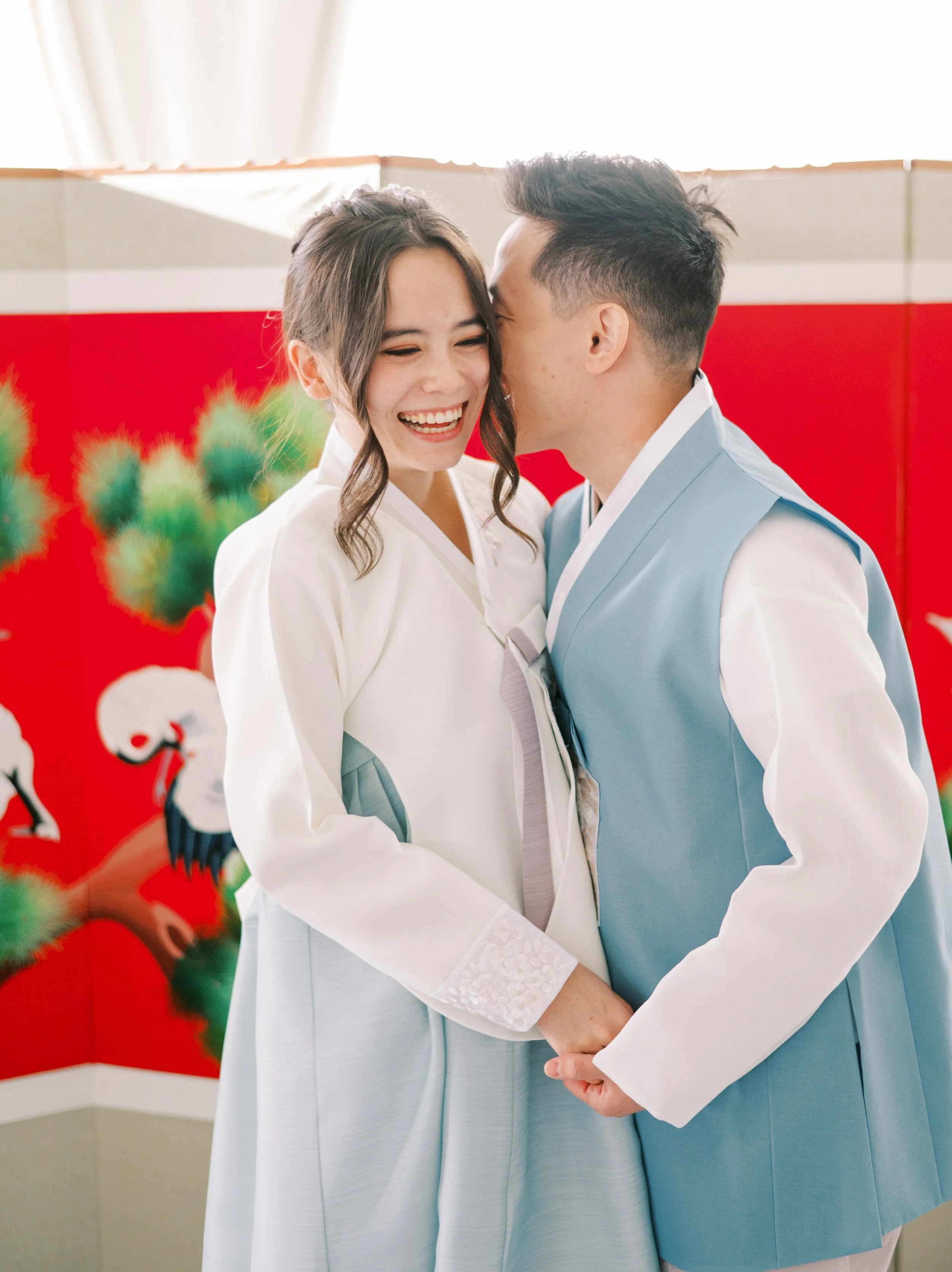 Bride and groom smile together during a Korean tea ceremony in front of a red traditional backdrop at The Ivory Hall, blending heritage and modern elegance.