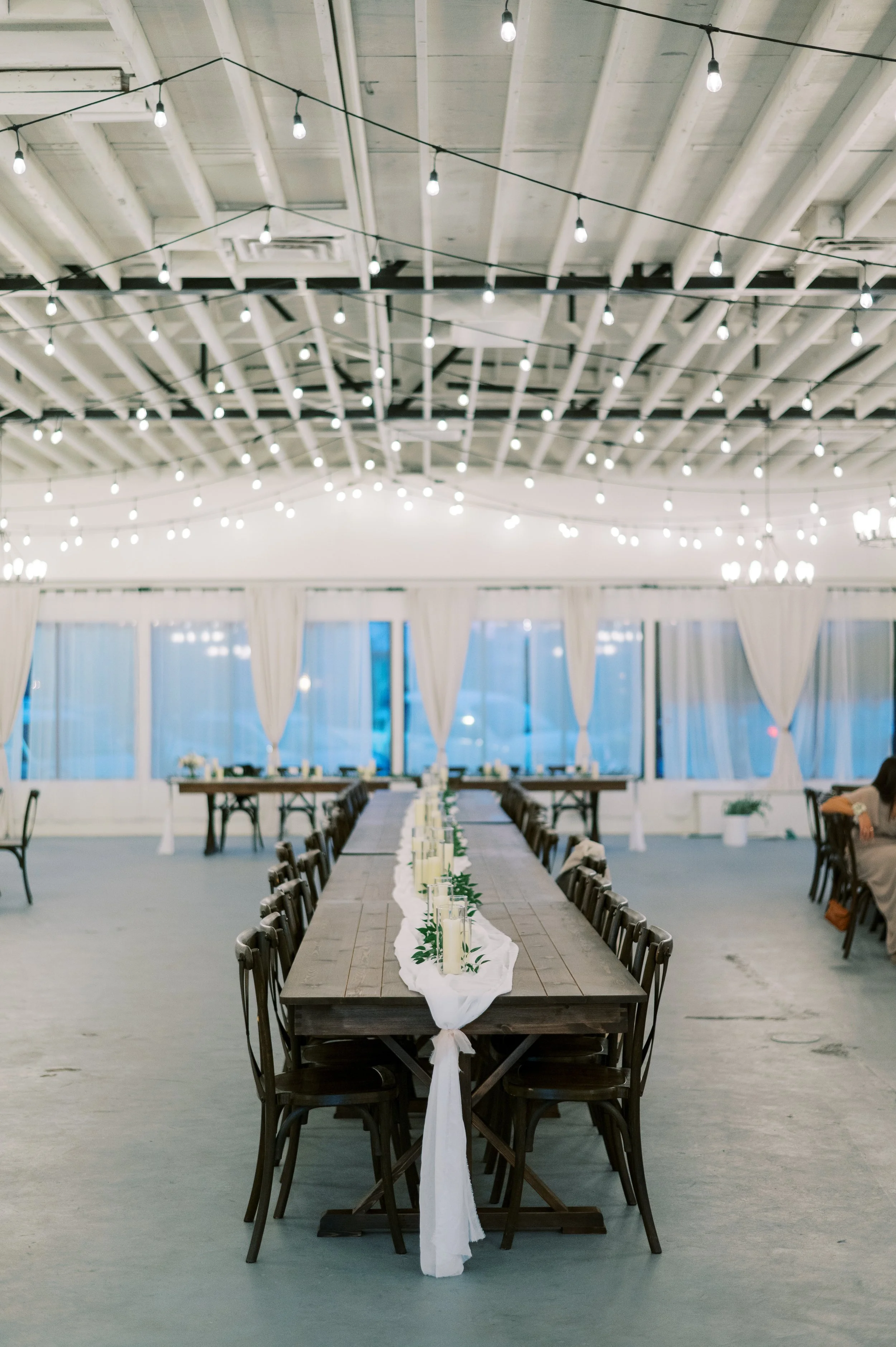 Reception tables at The Ivory Hall set for an elegant evening wedding, with long wood tables, simple greenery runners, and glowing candlelight under string lights and natural white drapes.