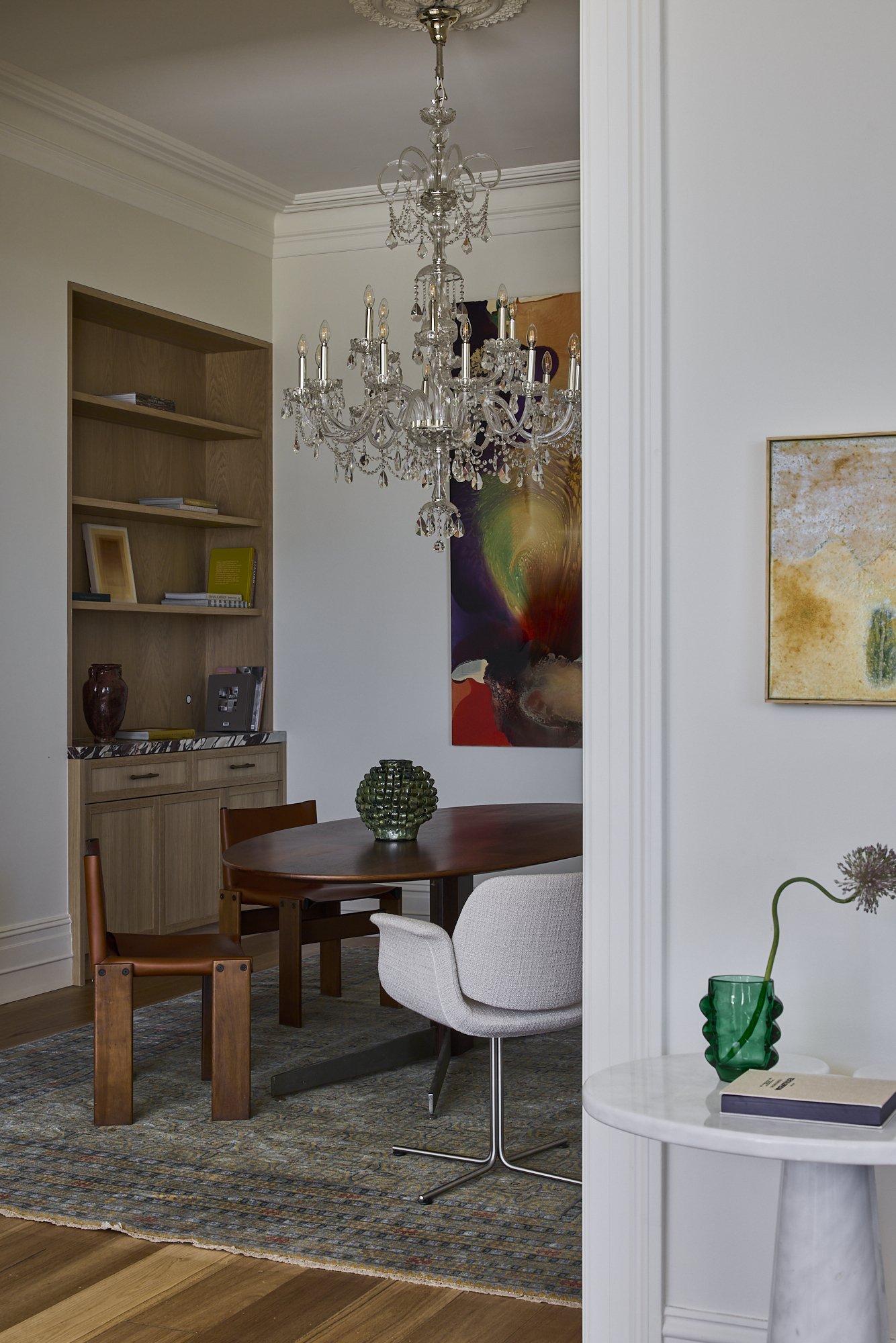 A dining room with a crystal chandelier, a wooden oval table, and a mix of chairs including an upholstered white chair, a brown wooden chair, and a small side table with a green vase and a book.
