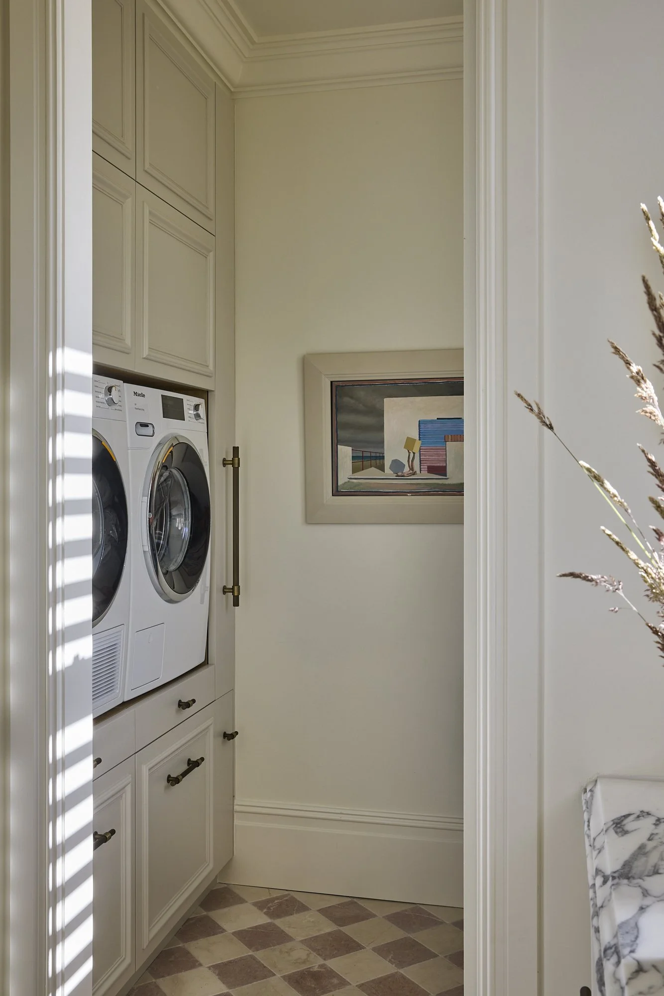 Laundry room with stacked front-loading washer and dryer, white cabinets above, framed artwork on the wall, and a partial view of a marble countertop.
