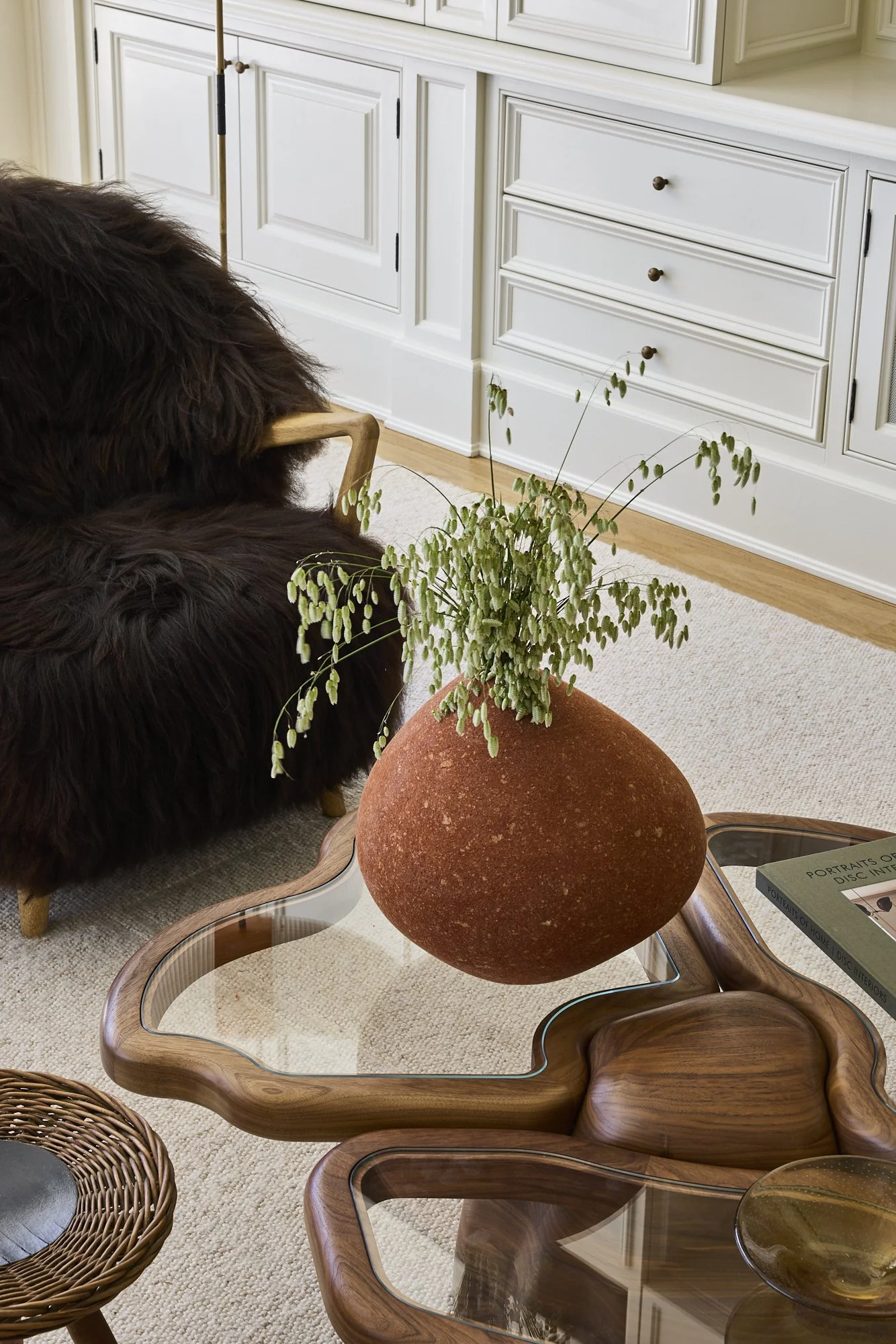 A round, red, textured vase with a green, leafy plant on a glass and wood coffee table in a living room with white cabinets and a beige rug.